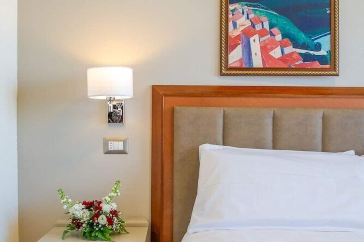 Une chambre d’hôtel avec un lit, une table de chevet avec une lampe, un tableau encadré et un arrangement floral.