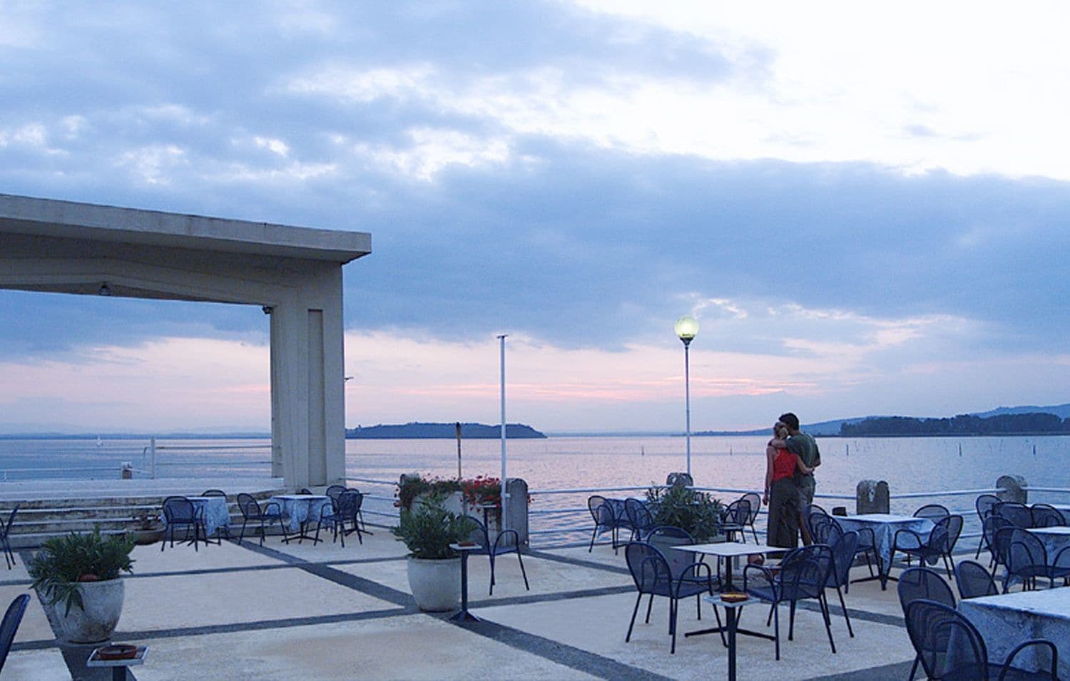 An outdoor seating area with tables and chairs, overlooking a body of water with distant landforms.