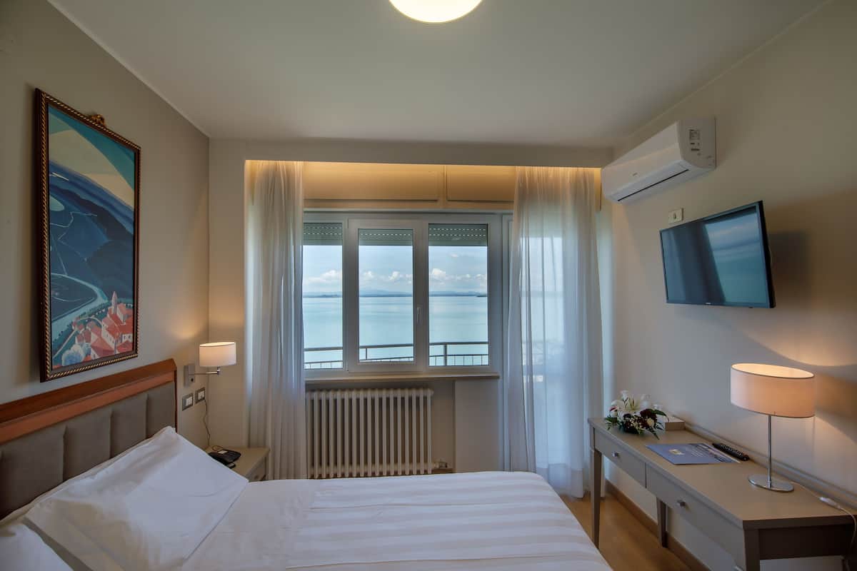 Ett hotellrum med en säng, ett skrivbord, en TV, ett fönster med utsikt över havet och en luftkonditionering.