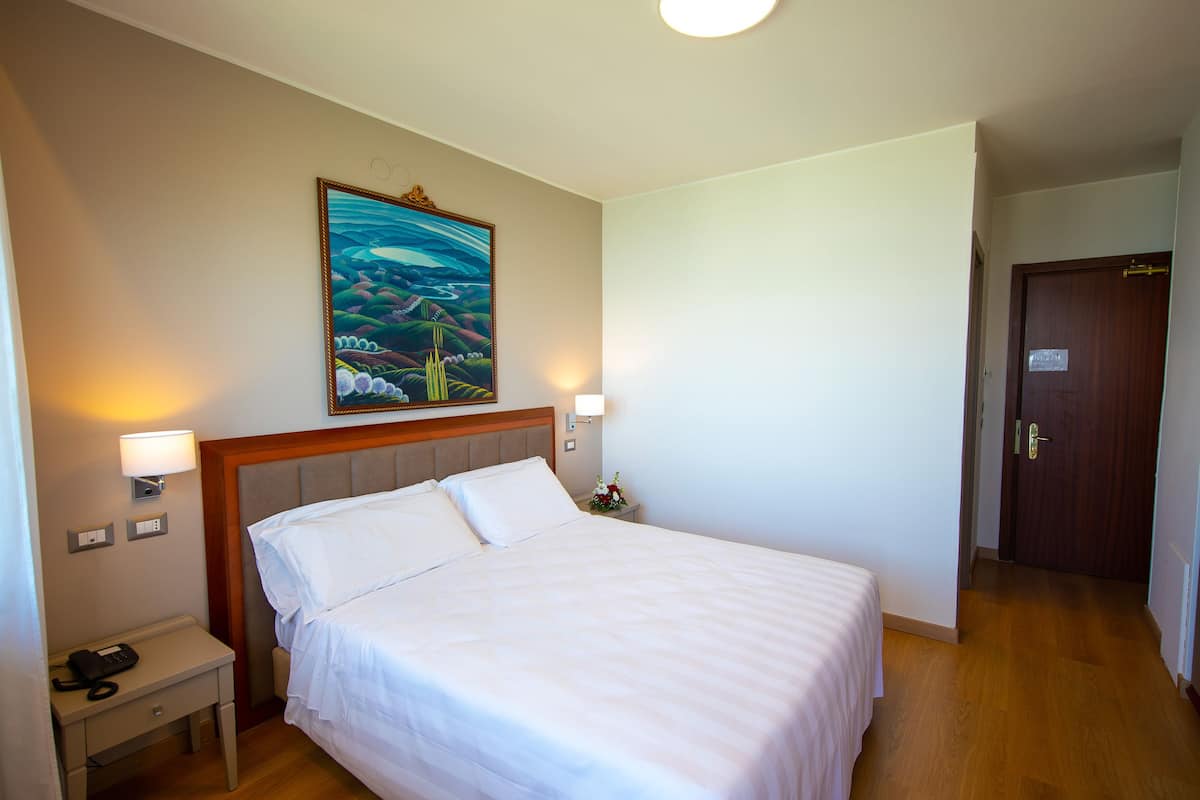 Ett hotellrum med en stor säng, två sänglampor, ett nattduksbord med en telefon, en tavla på väggen och en dörr av trä.