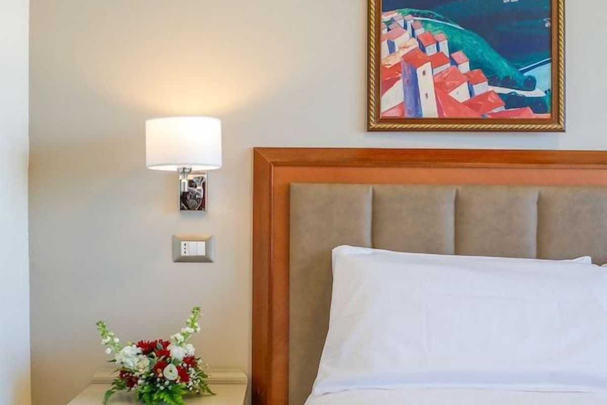 A hotel room with a bed, a nightstand with a lamp, a framed painting, and a floral arrangement.