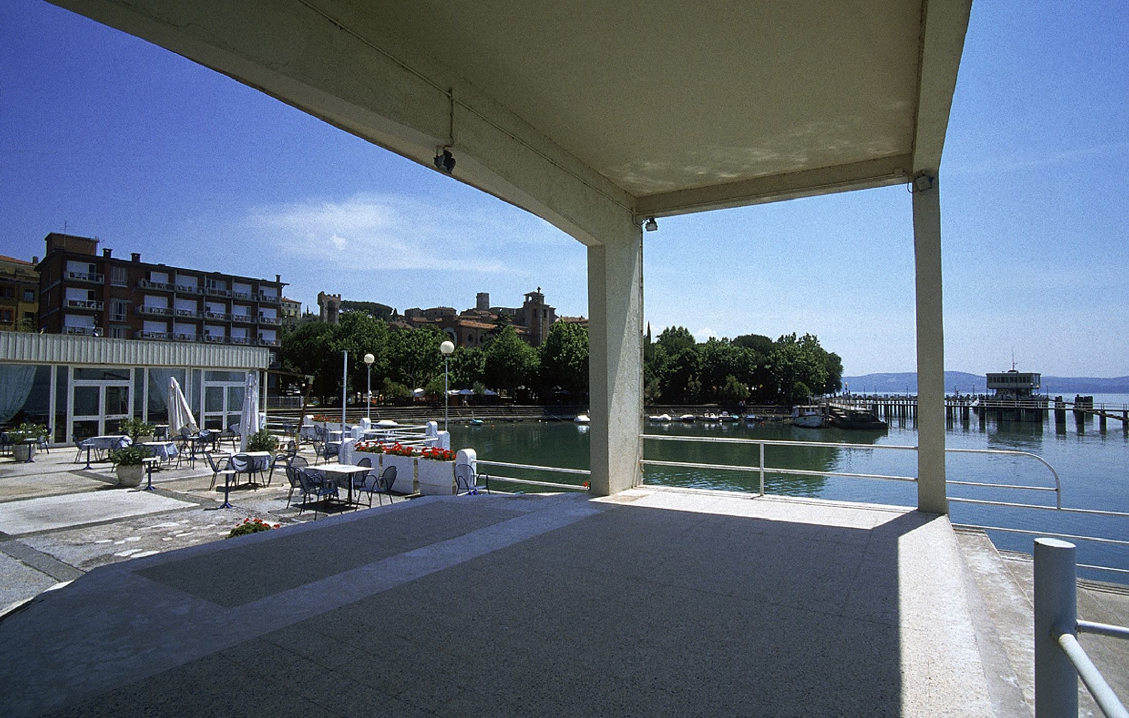 Un espacio al aire libre con mesas y sillas, con vista a un cuerpo de agua con barcos y un muelle.