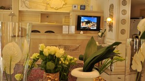 A hotel reception area with a television displaying a cityscape, a reception desk, and various floral arrangements. - Hotel Jolanda (Santa Margherita Ligure)