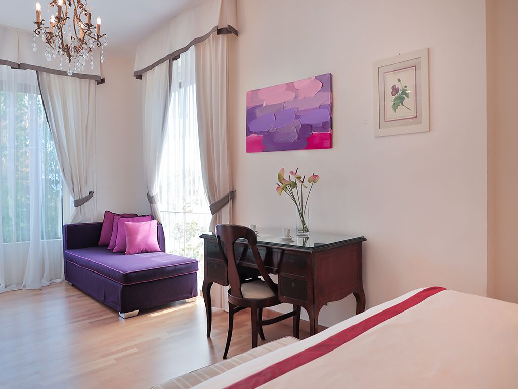 A room with a bed, a desk, a chair, a sofa, a chandelier, and artwork on the walls.