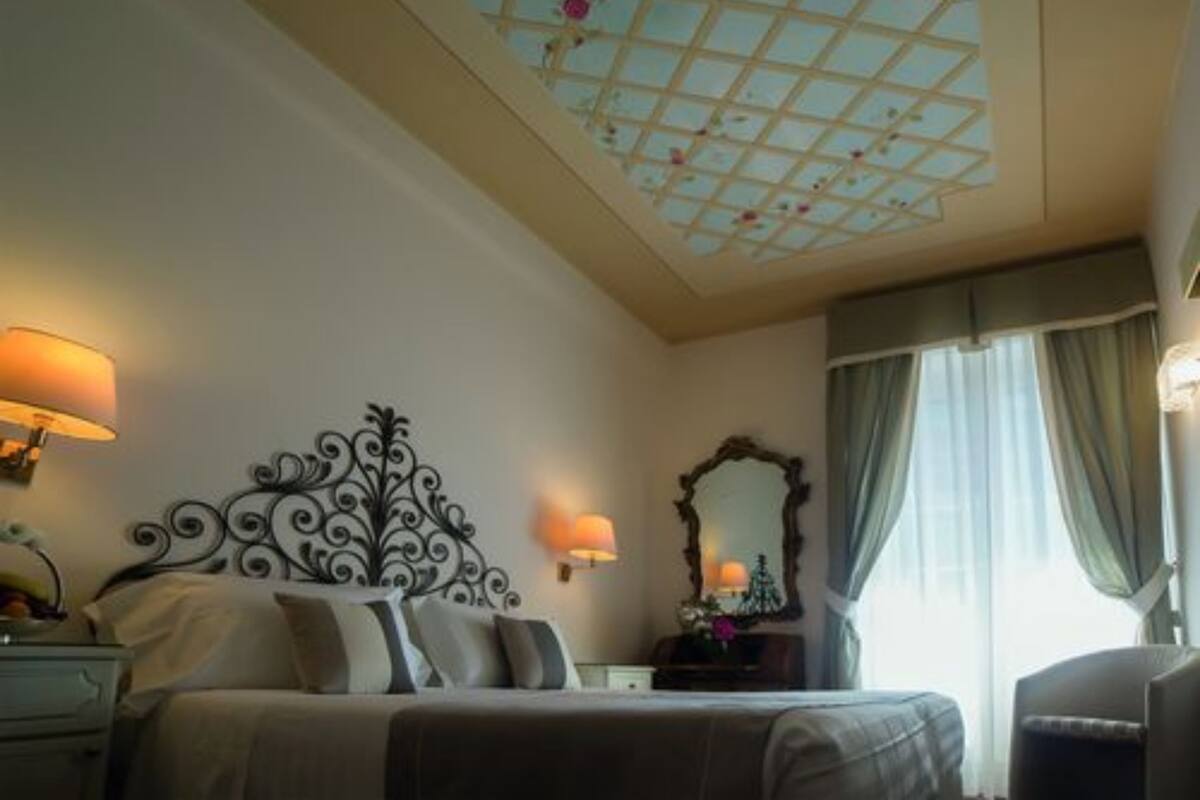 Une chambre d’hôtel avec un lit, une table de chevet, une lampe, un miroir et une fenêtre avec des rideaux.