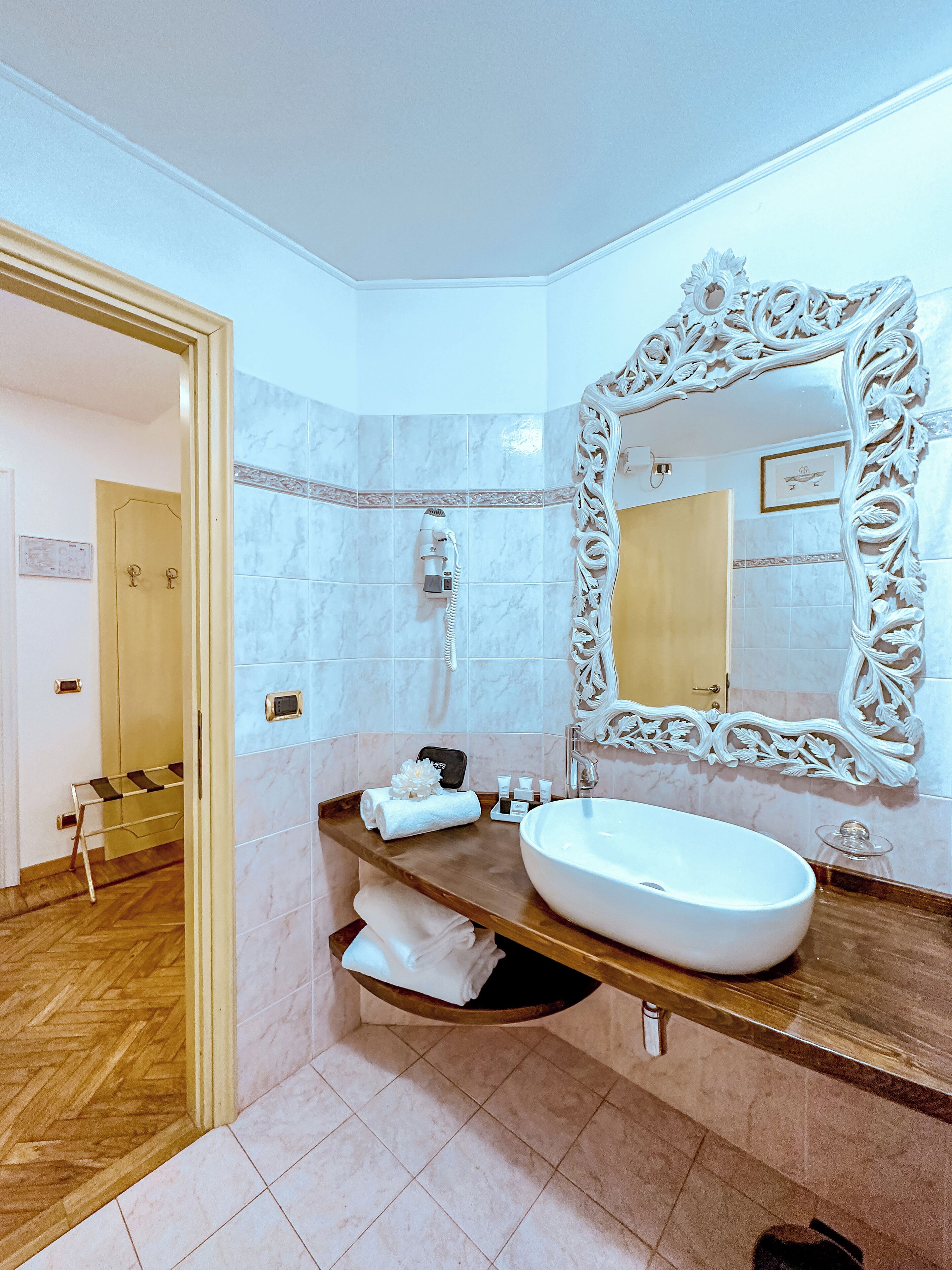 A bathroom with a large ornate mirror, a wooden vanity, a white sink, and a towel rack.