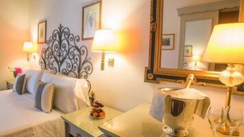 A hotel room with a bed, bedside table, lamp, mirror, and a small table with a fruit bowl and wine glasses.