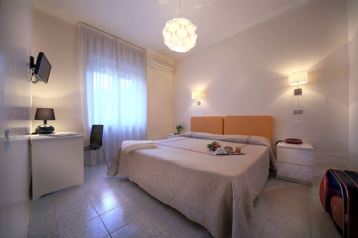 Hotellihuone, jossa on sänky, yöpöytä, lamppu ja matkalaukku.