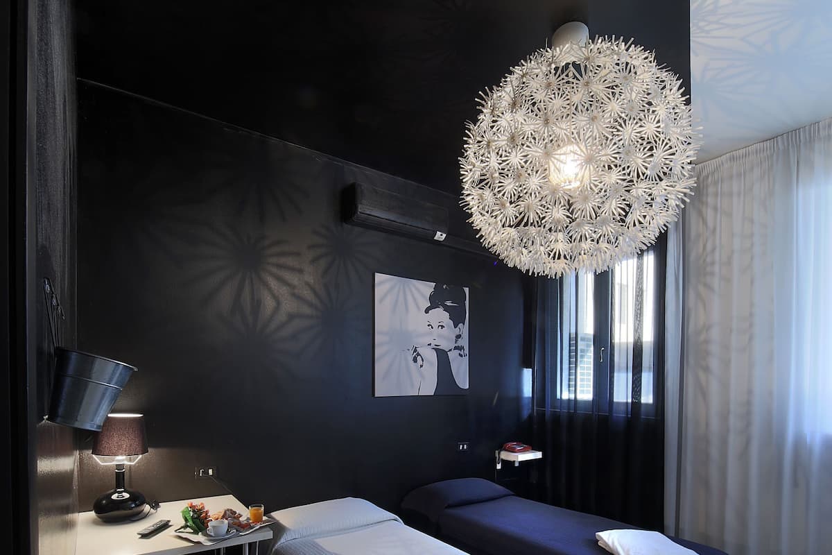 Moderni hotellihuone, jossa on tummat värit, suuri koristeellinen valaisin, sänky, yöpöytä lampun kanssa ja pieni valkoinen tuoli.