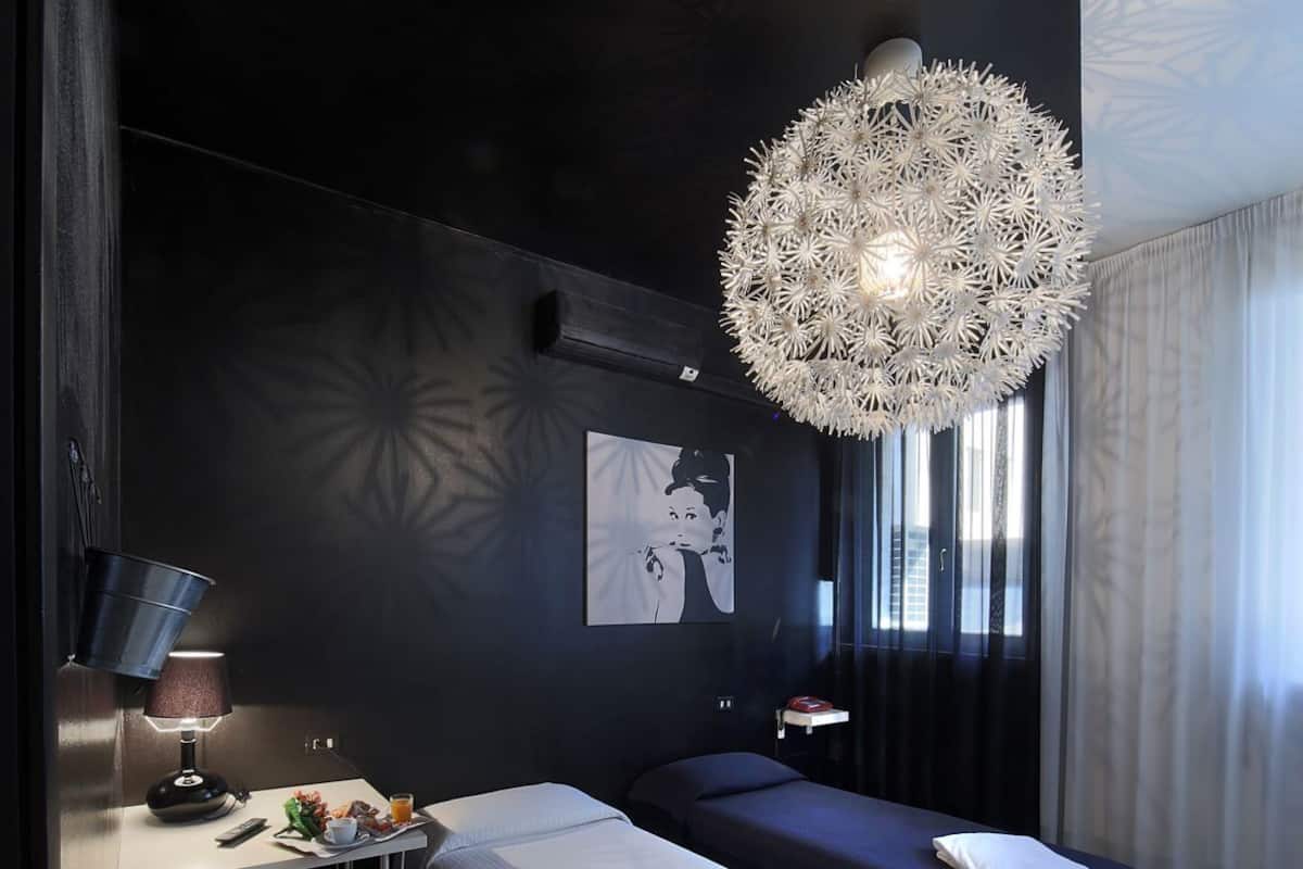 Moderni hotellihuone, jossa on tummat värit, ainutlaatuinen kattokruunu, sänky, yöpöytä lampun kanssa ja seinälle kiinnitetty taideteos.