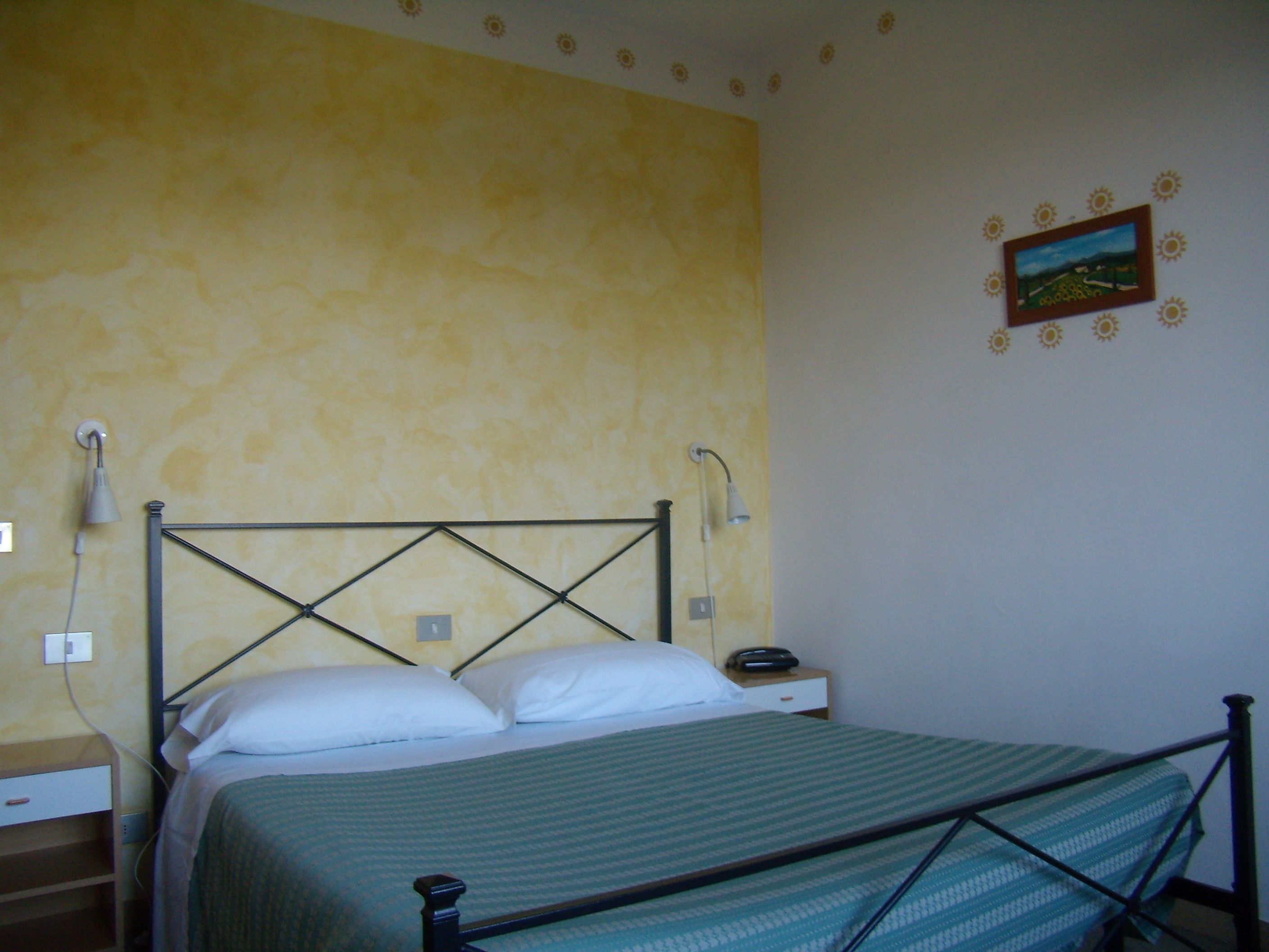 A single bed with a metal frame, a bedside table with a lamp, and a wall with a floral pattern and a framed picture.