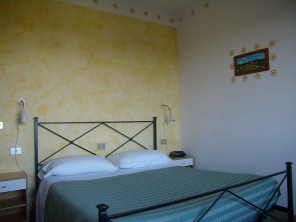 A single bed with a metal frame, a bedside table with a lamp, and a wall with a floral pattern and a framed picture.