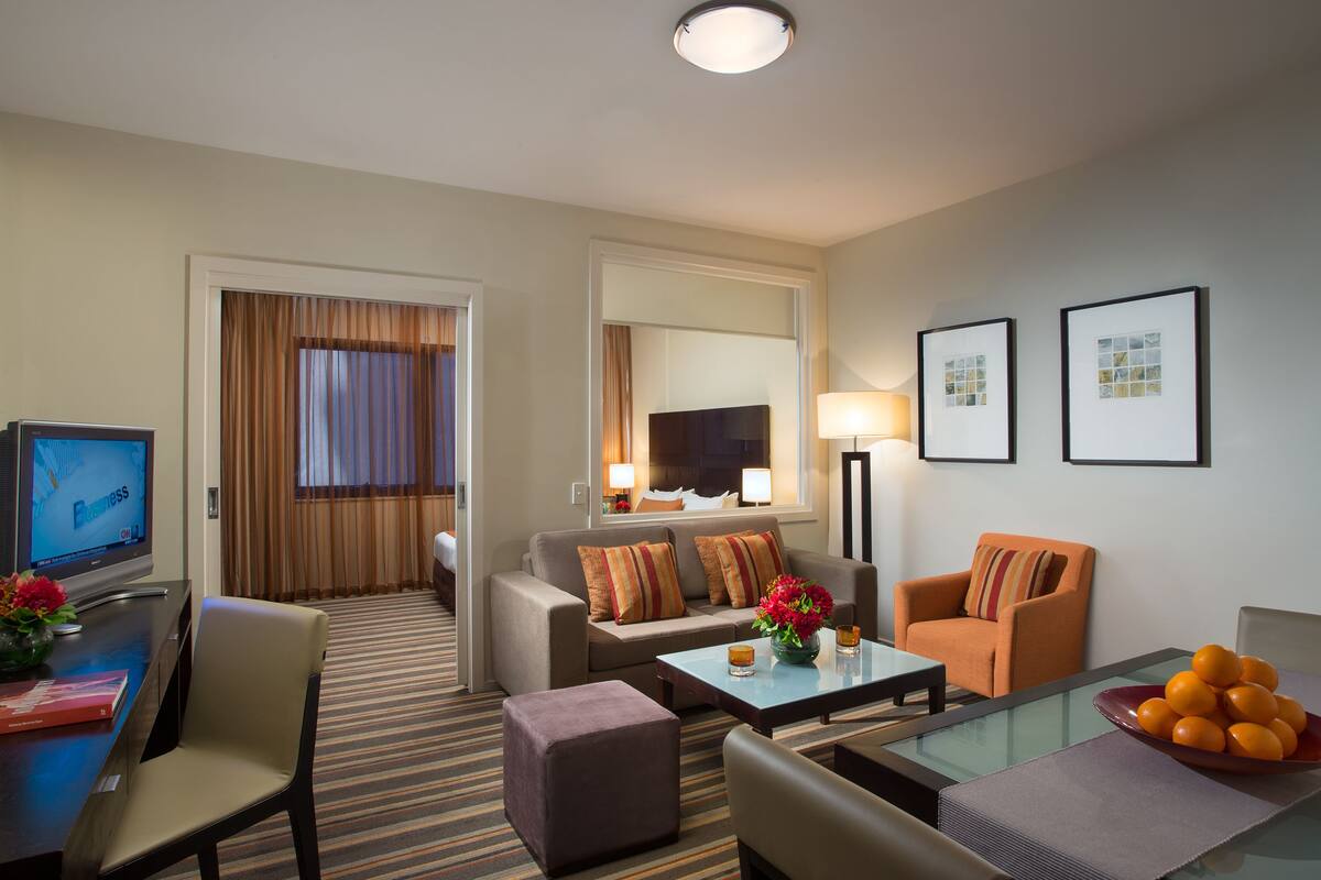 A hotel room with a living area, a television, a sofa, a coffee table, a dining table with a fruit bowl, and a flat-screen TV.