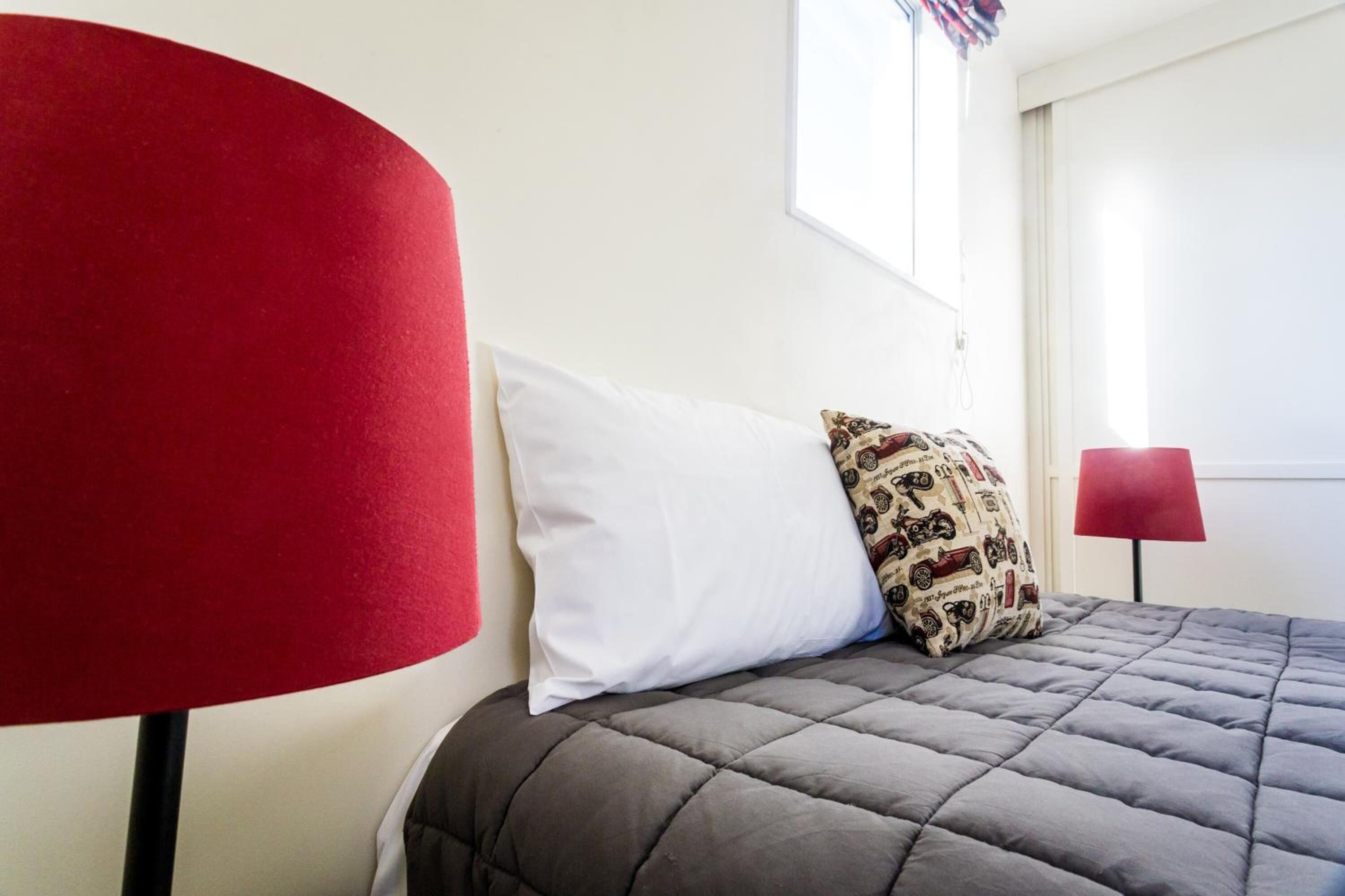 Una cama con una colcha gris acolchada, una almohada blanca y una almohada estampada. A la izquierda de la cama hay una lámpara roja.