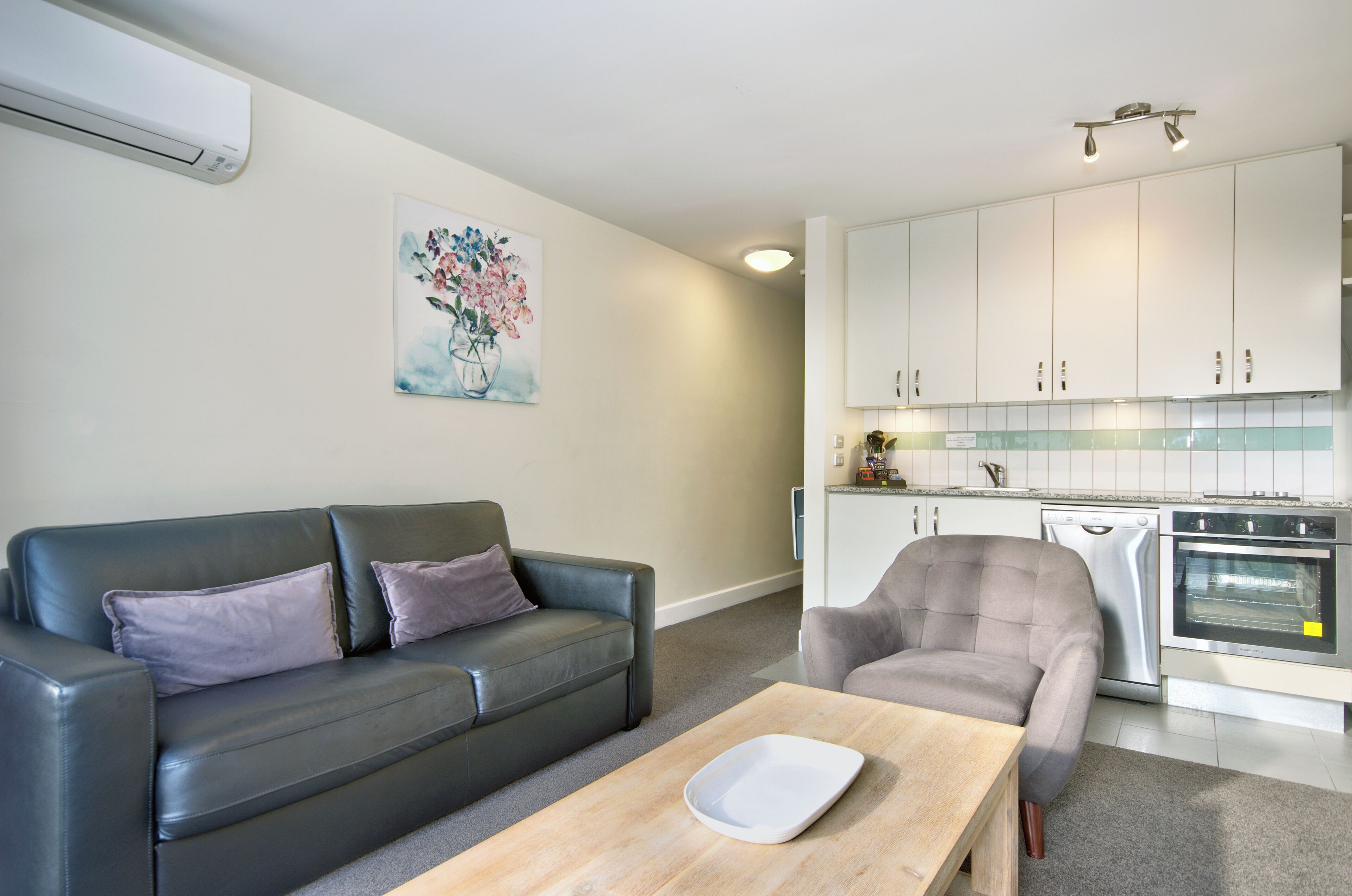 A modern living room with a grey leather sofa, a wooden coffee table, and a grey armchair. A kitchen with white cabinets and stainless steel appliances is visible in the background.