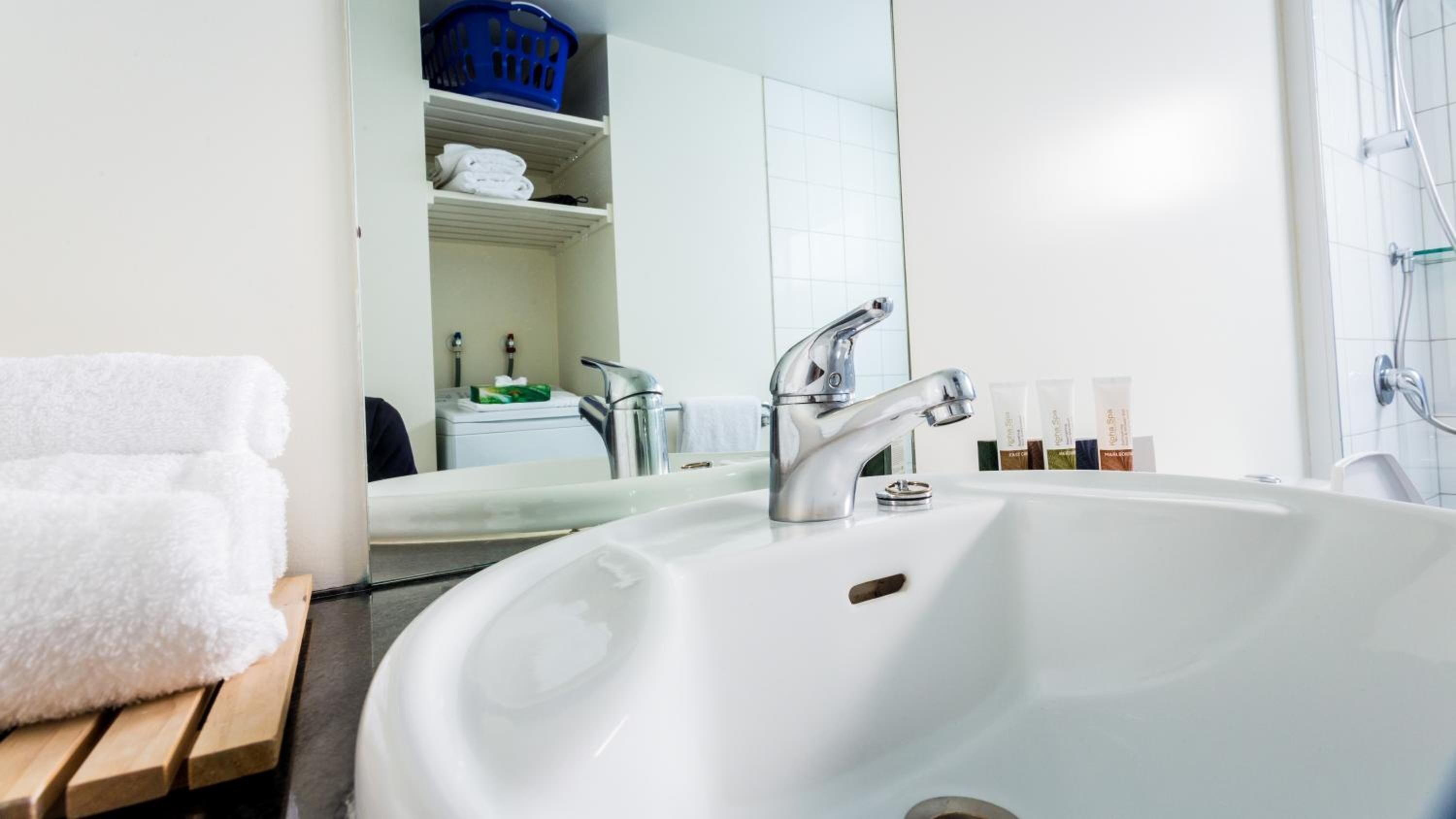 A bathroom with a white sink, two faucets, a mirror, and a blue laundry basket on the shelf.