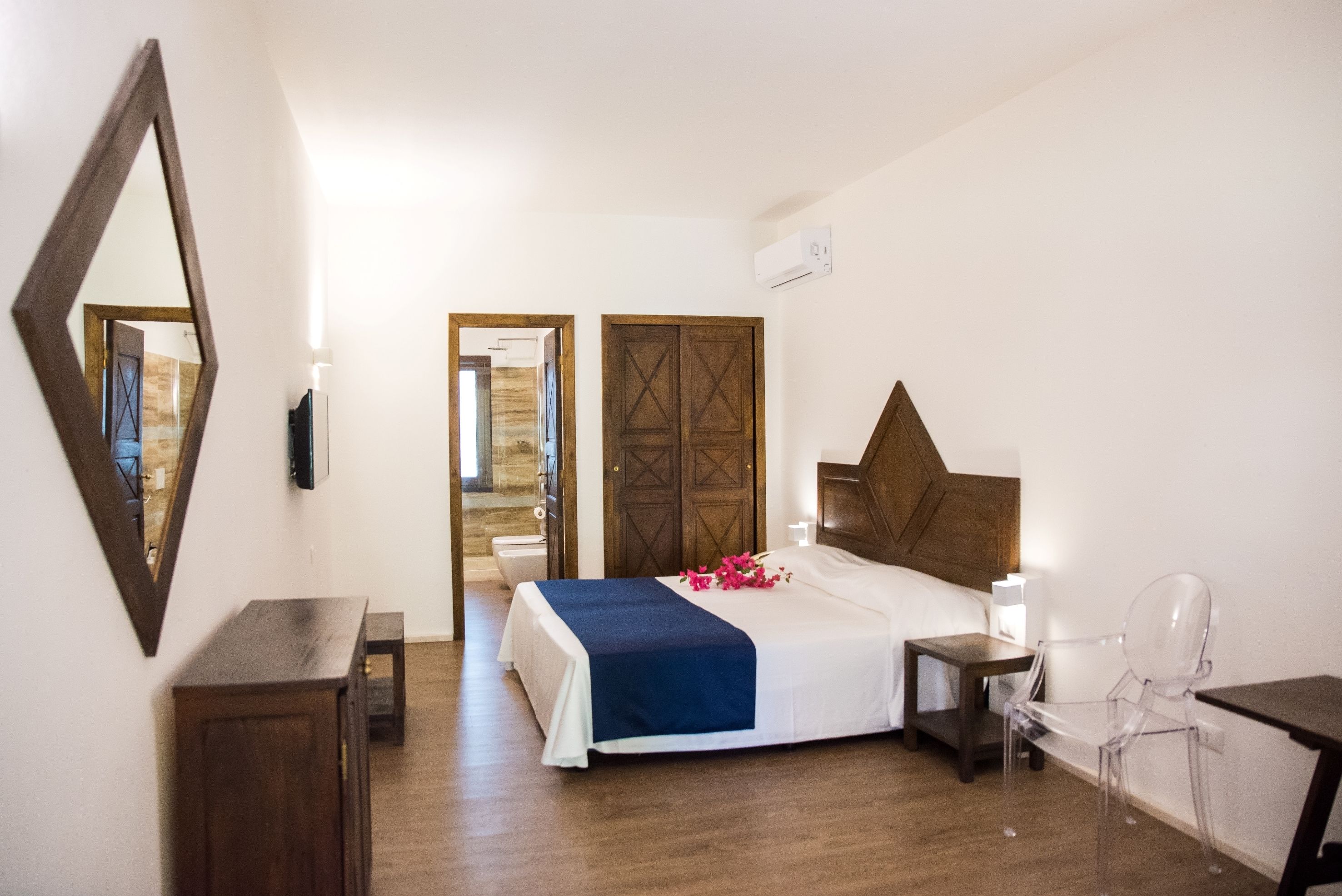 A hotel room with a bed, bedside table, chair, wooden wardrobe, and a mirror.