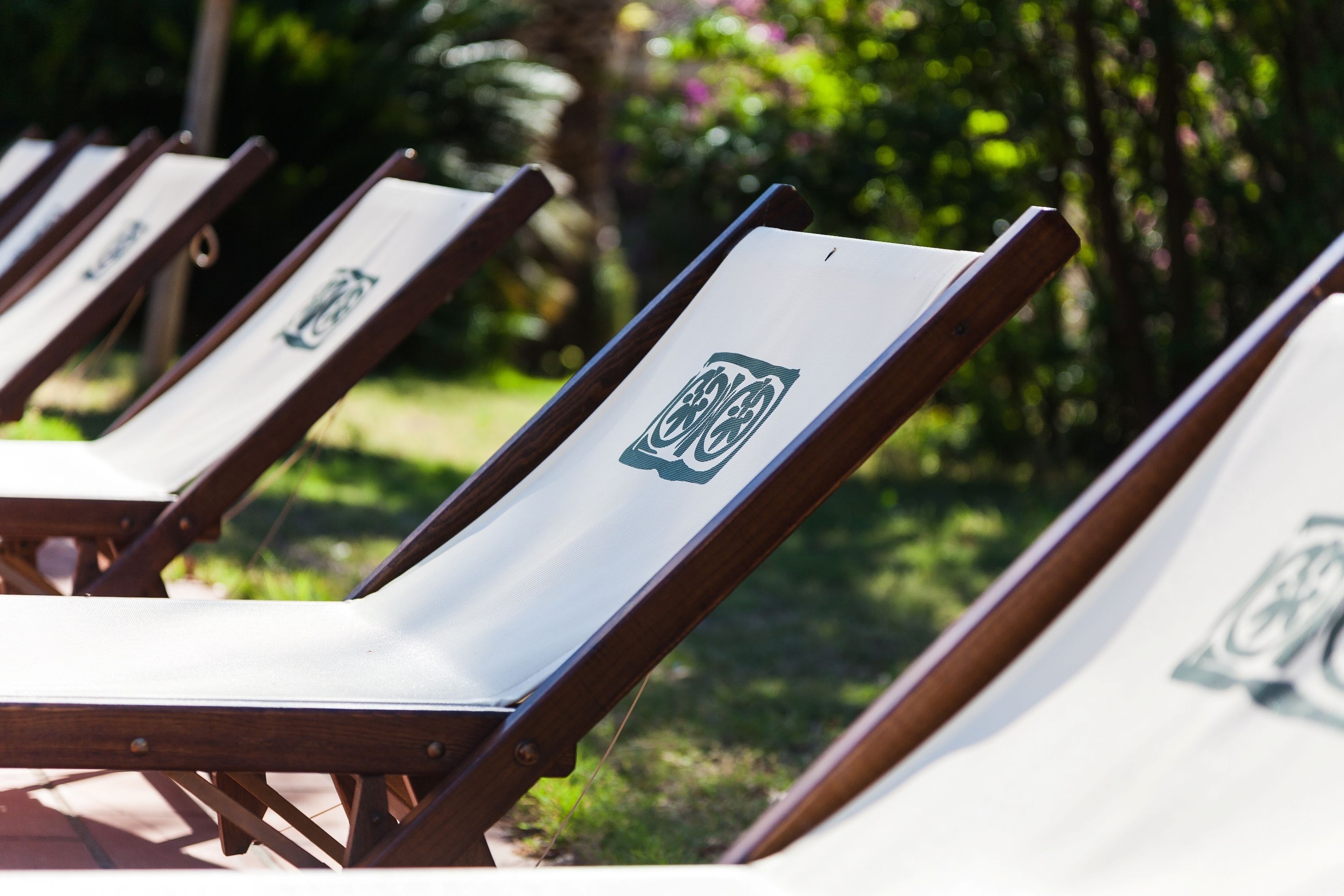 A row of wooden lounge chairs with white cushions and a logo on the backrest, set against a lush green garden backdrop.