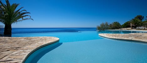 A clear blue pool with a curved edge, surrounded by stone flooring and palm trees, leading to a beach and ocean view.