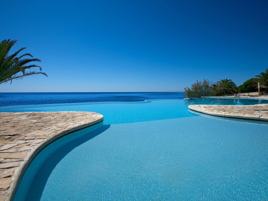 Piscina azul cristalina con borde curvo, rodeada de pavimento de piedra y palmeras, que da a una playa y vista al océano.