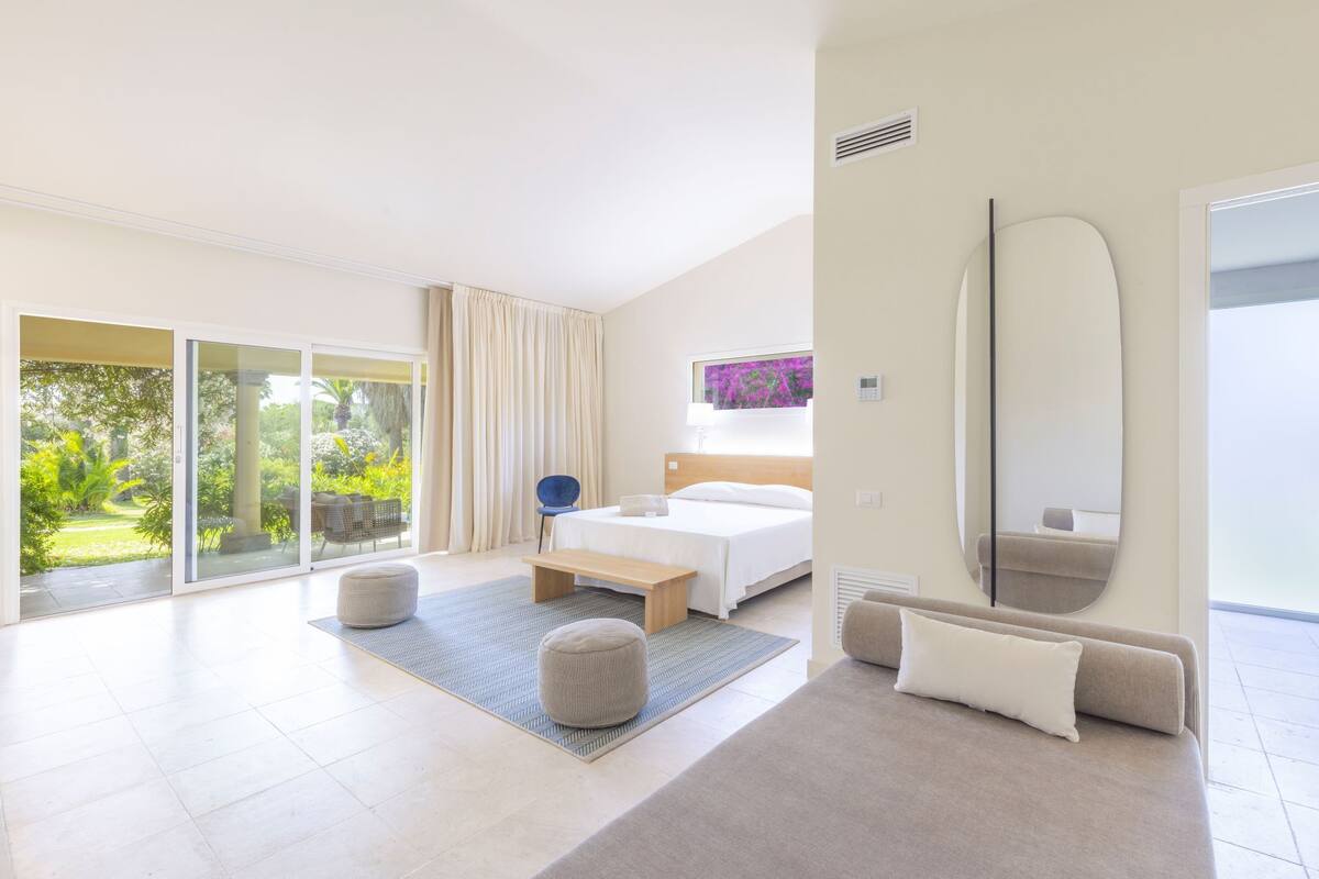 Een moderne slaapkamer met een groot bed, een bankje, poefs, een bank en een grote spiegel.