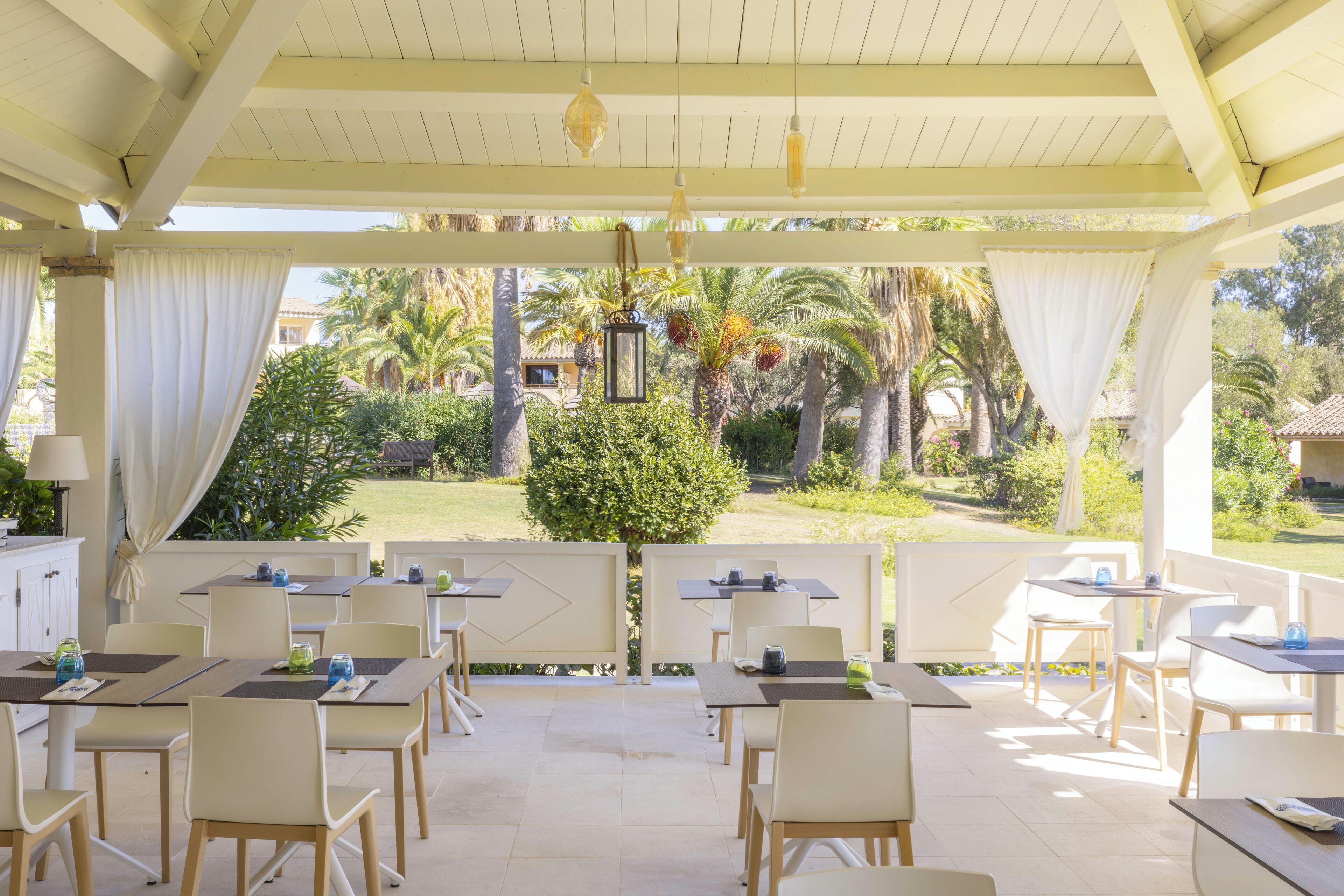 An outdoor patio with tables and chairs, a pergola with hanging lights, and a view of a garden with palm trees and a small tree in the center.