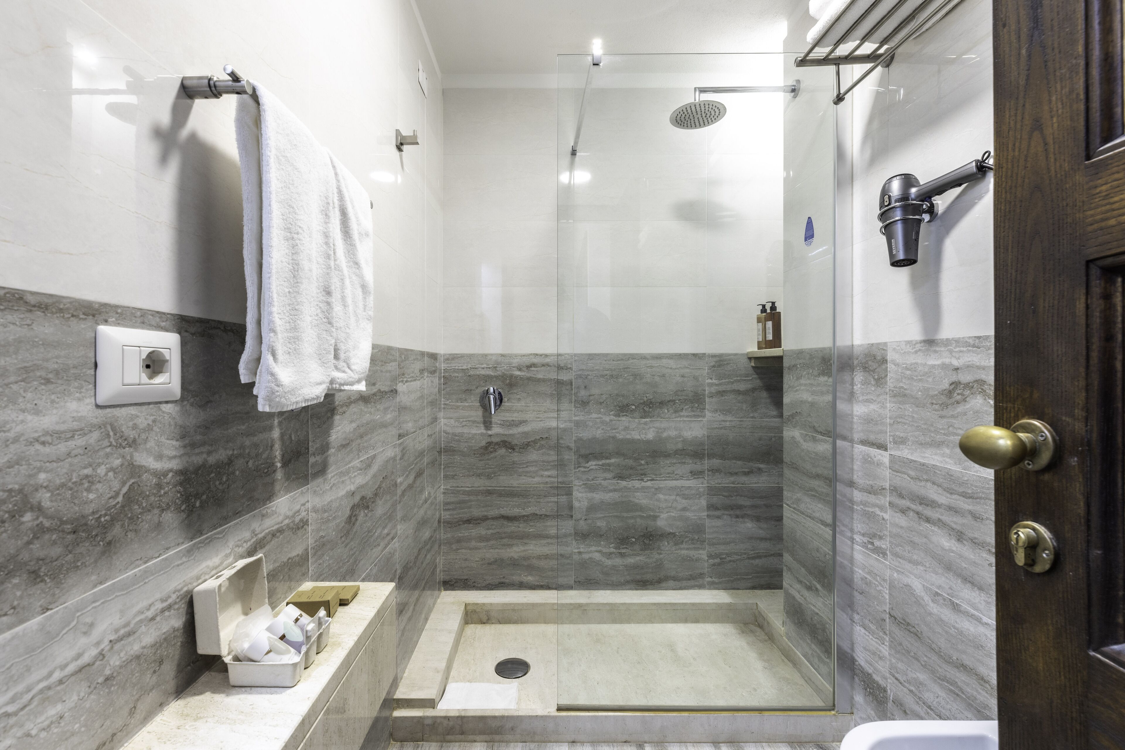 A modern bathroom with a glass-enclosed shower, marble walls, a towel rack, and a small sink area with toiletries.