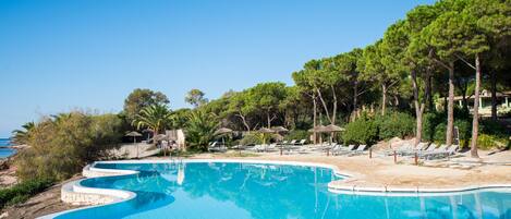 A clear blue pool with a curved edge, surrounded by lounge chairs and umbrellas, with a beach and trees in the background.