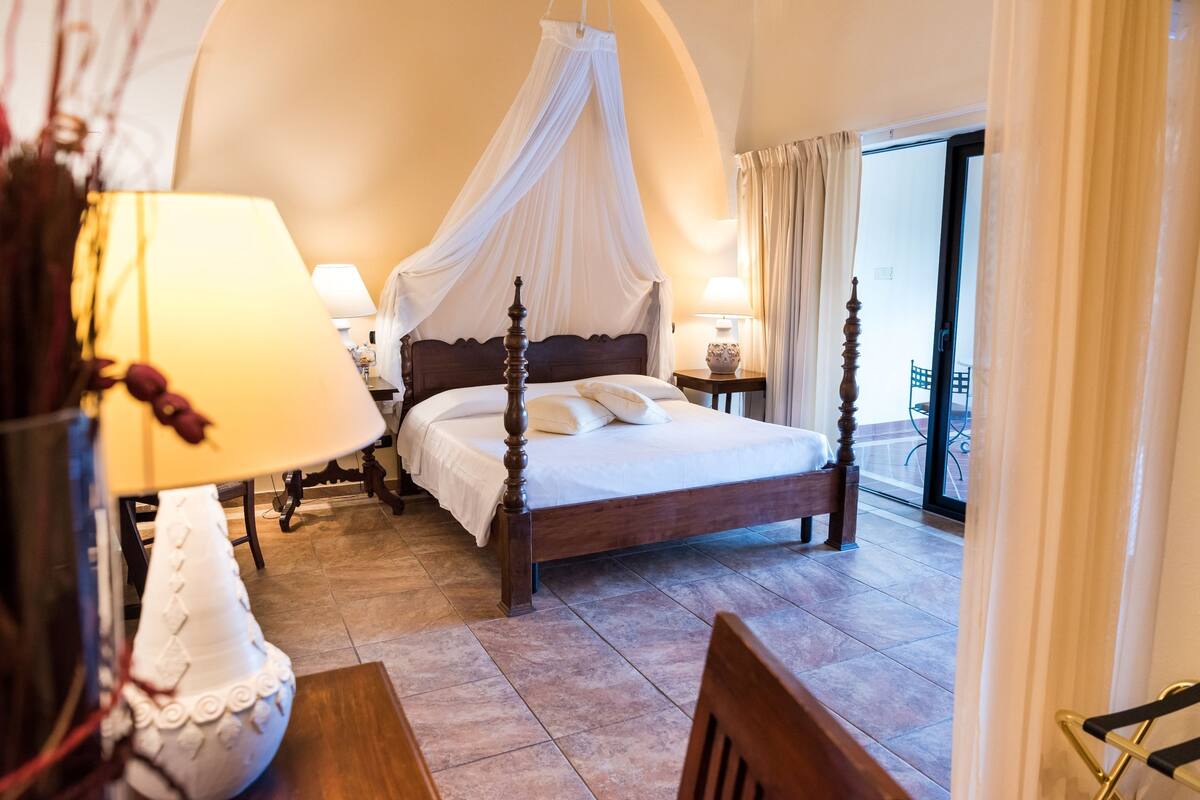 Een slaapkamer met een hemelbed, nachtkastje, lamp en uitzicht op het balkon.