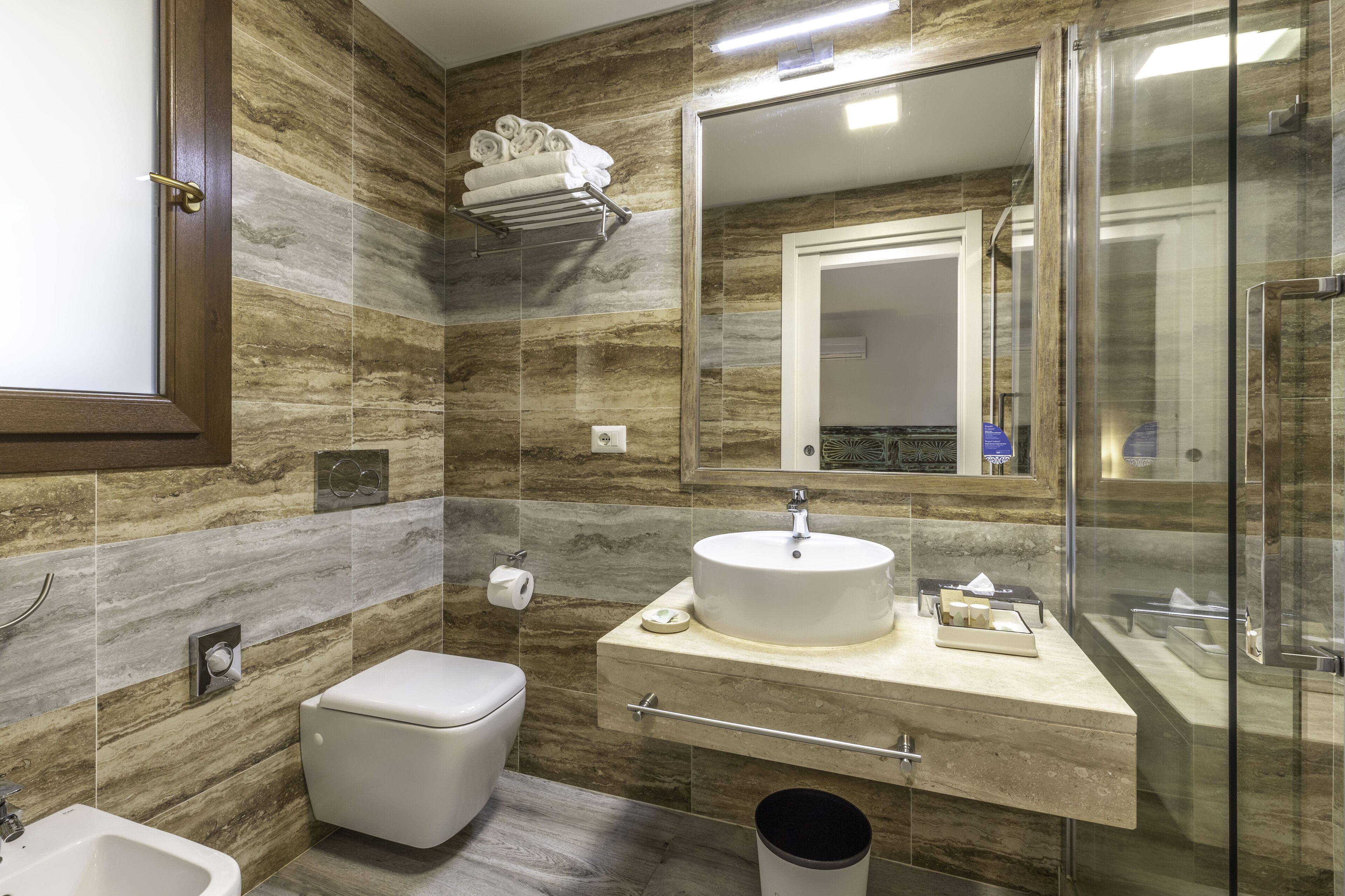 A modern bathroom with a marble backsplash, a glass shower enclosure, a white sink, and a toilet.