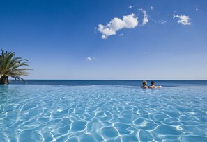 A clear blue pool with two people in the water, surrounded by a palm tree and a view of the ocean. - Hotel Costa Dei Fiori (Pula)