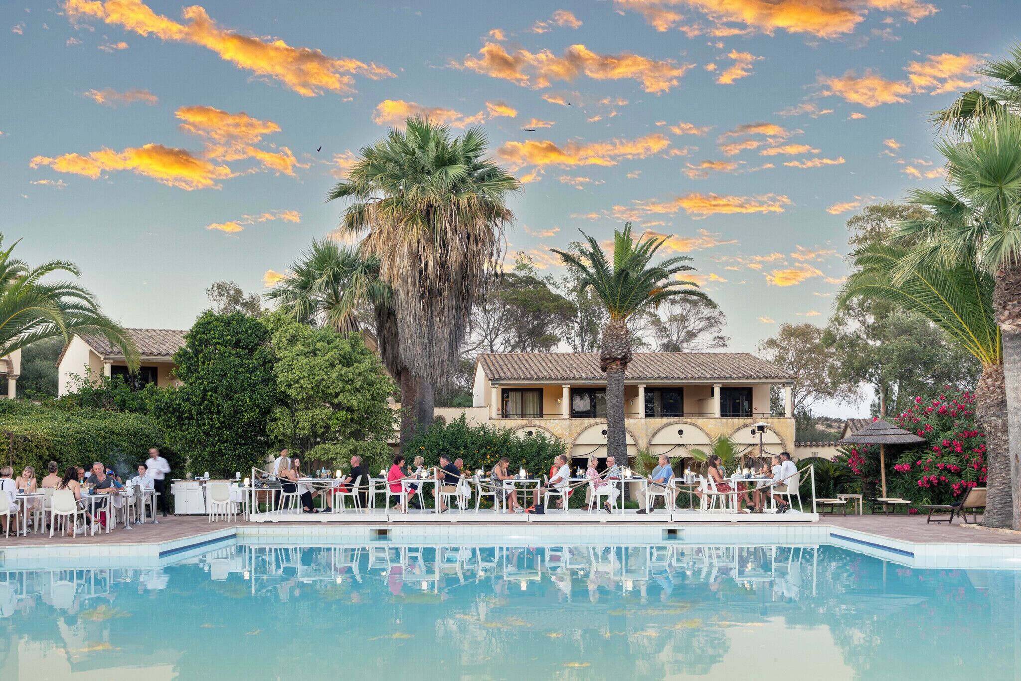 A poolside area with tables and chairs, palm trees, and a building in the background.