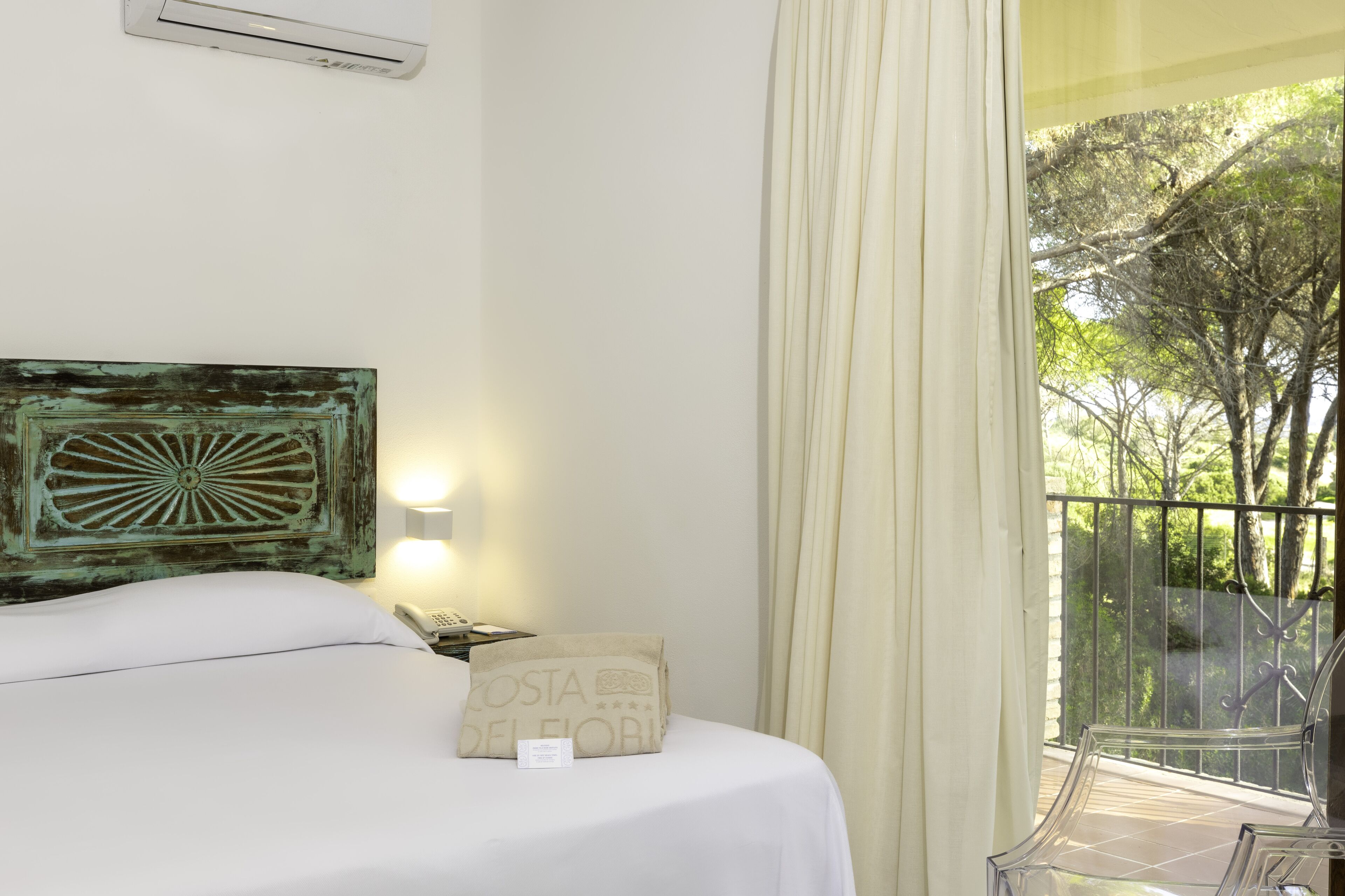 A hotel room with a bed, a nightstand, a lamp, a decorative wall piece, a window with curtains, and a view of trees.