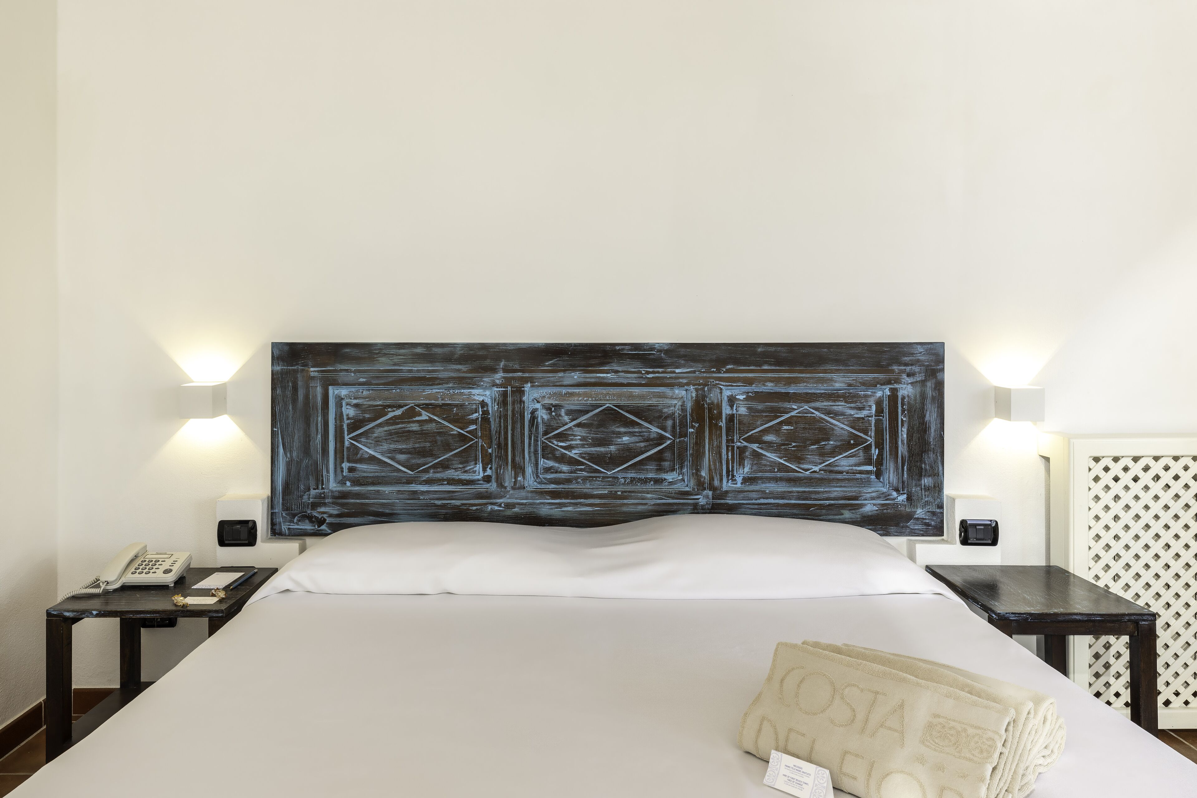 A neatly made bed with a white sheet, a dark headboard with geometric patterns, bedside lamps, a nightstand with a phone and a notepad, and a folded towel with the word "COSTA" on it.