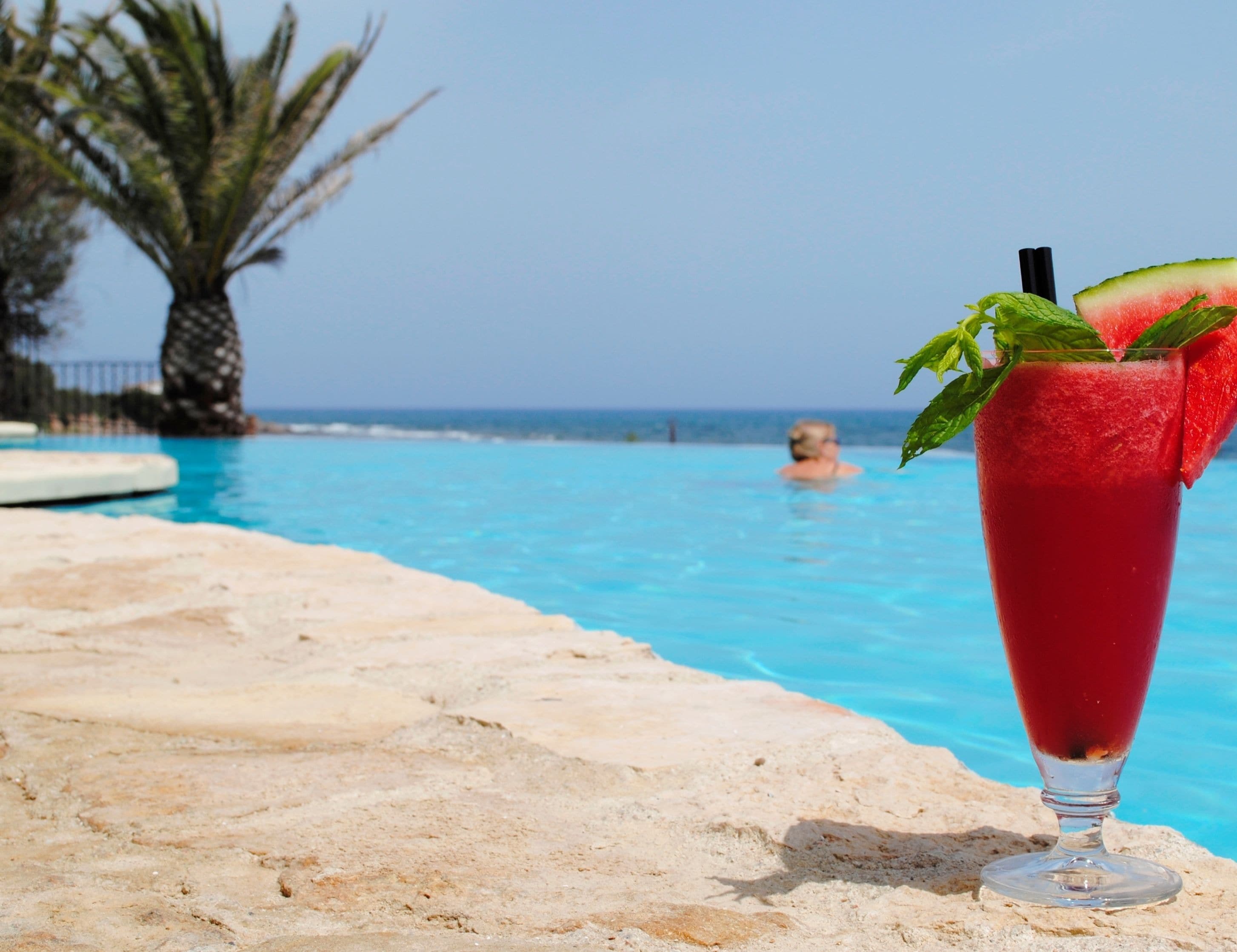 A clear pool with a glass of red drink on the edge, a palm tree, and a person in the background.