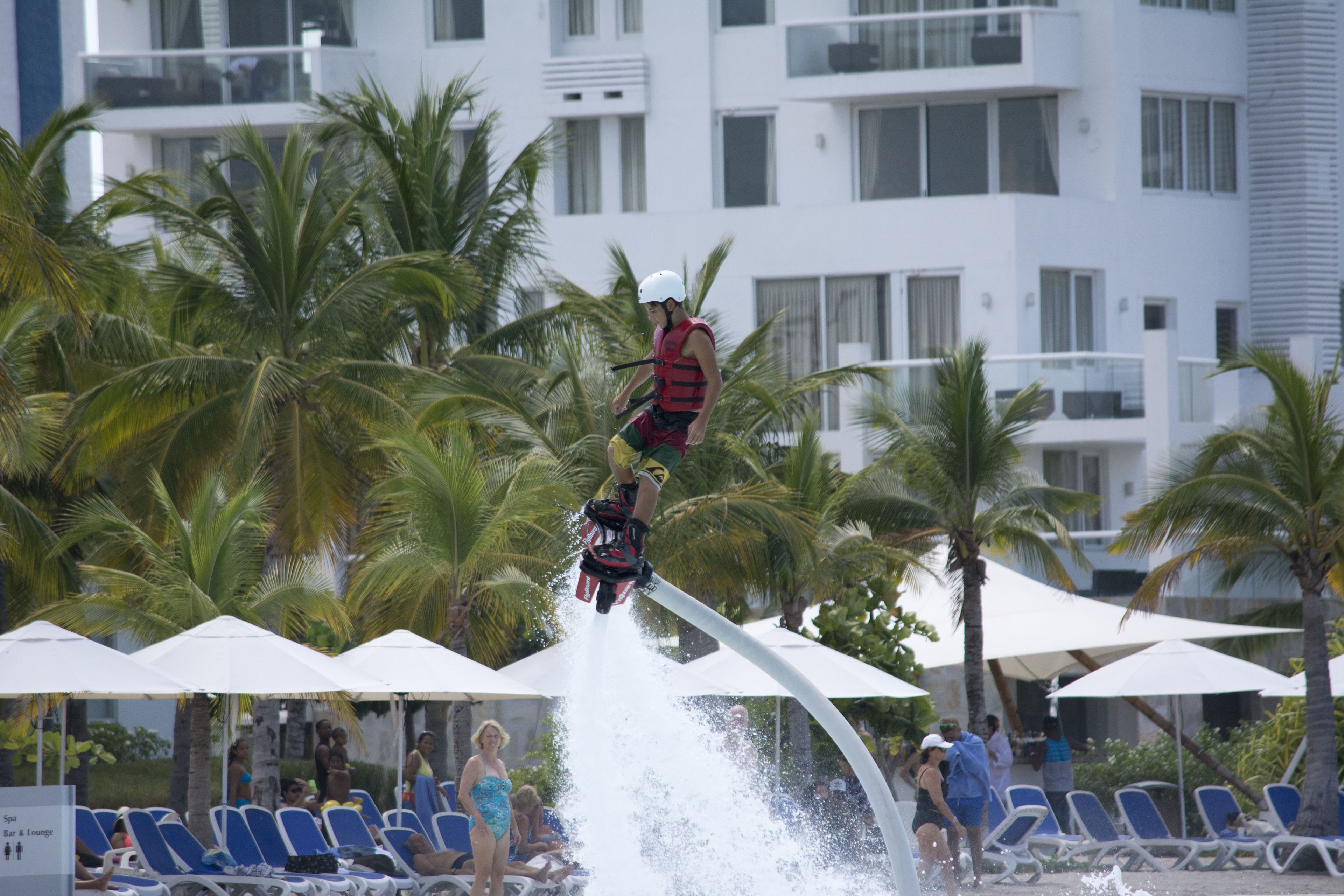 Una persona realiza una acrobacia acuática en una zona de piscina con sillas de descanso y palmeras.