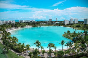 A large, clear blue pool surrounded by palm trees and a beach, with buildings in the background. - Playa Blanca Beach Resort - All Inclusive (Rio Hato)