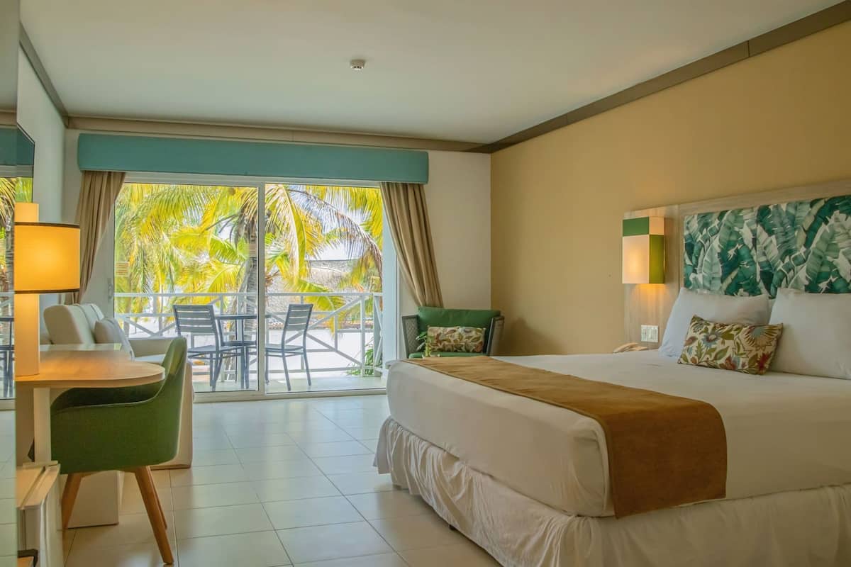 A hotel room with a bed, a desk, a chair, a balcony with a view of palm trees, and a tropical-themed headboard.