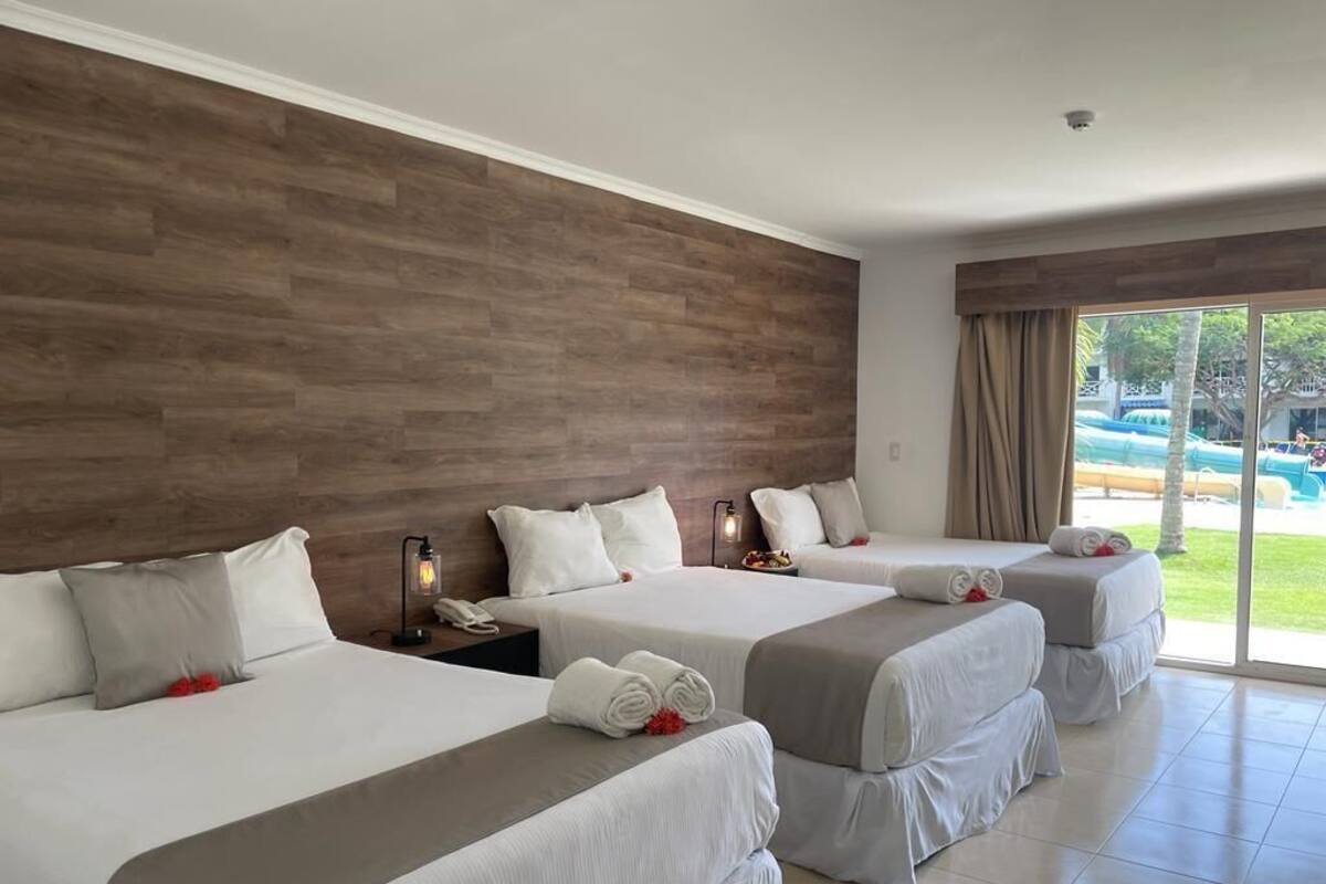 A hotel room with three beds, a wooden headboard, a large window with a view of a pool and greenery, and a nightstand with a lamp.
