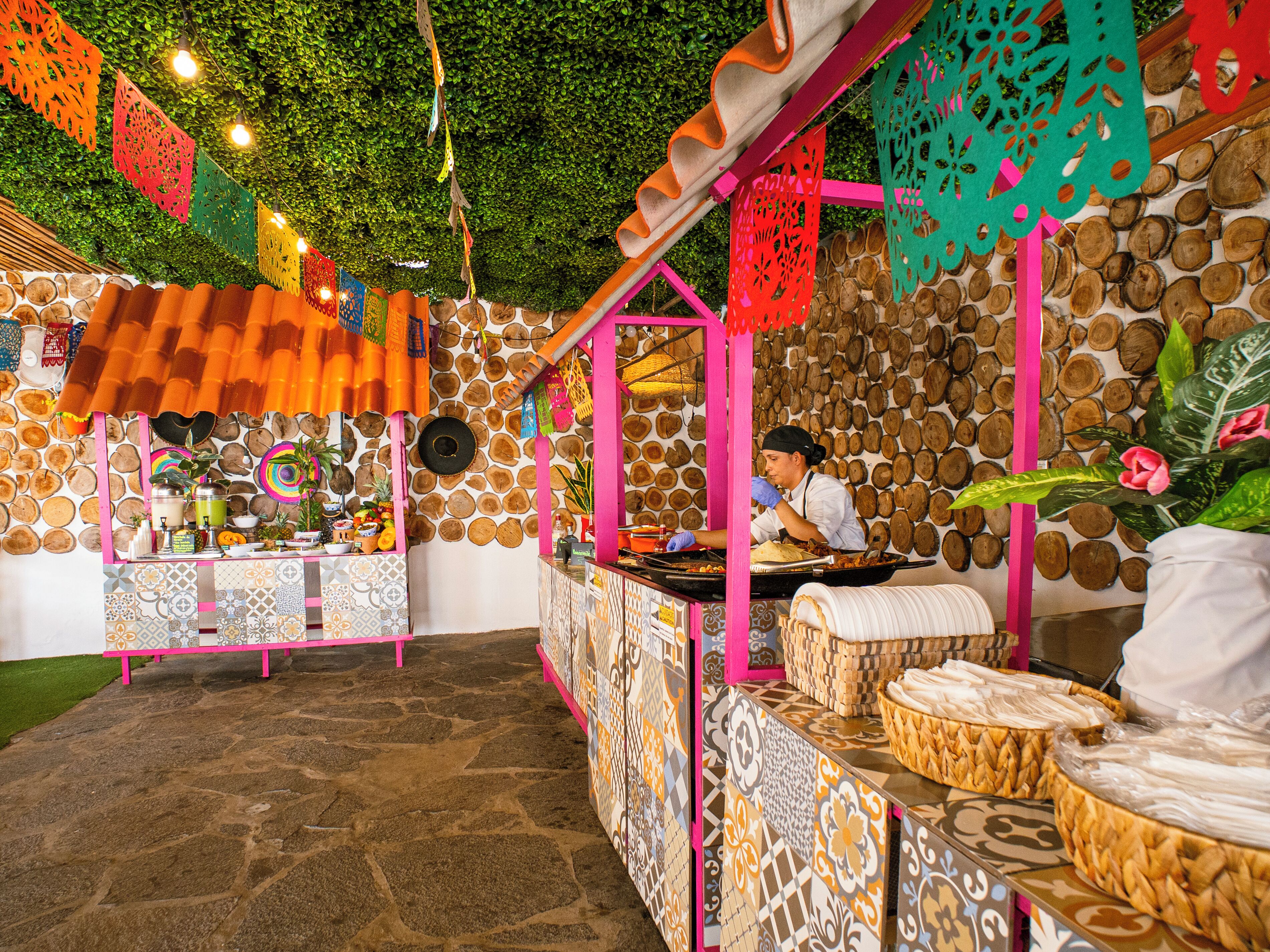 A vibrant outdoor dining area with a stone wall backdrop, colorful hanging decorations, and a chef preparing food.