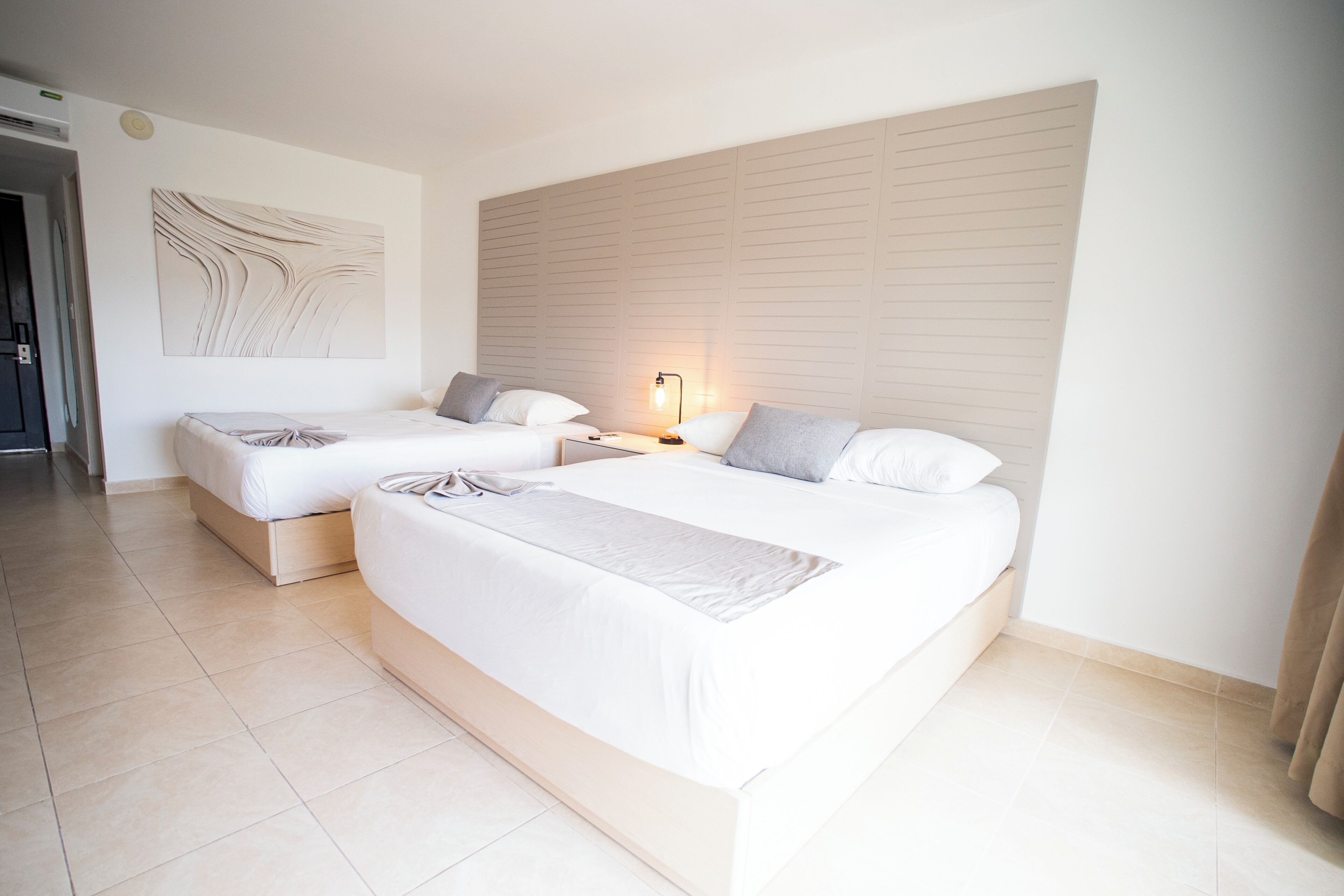 Habitación de hotel con dos camas, una obra de arte colgada en la pared, una lámpara de noche y piso de baldosas.