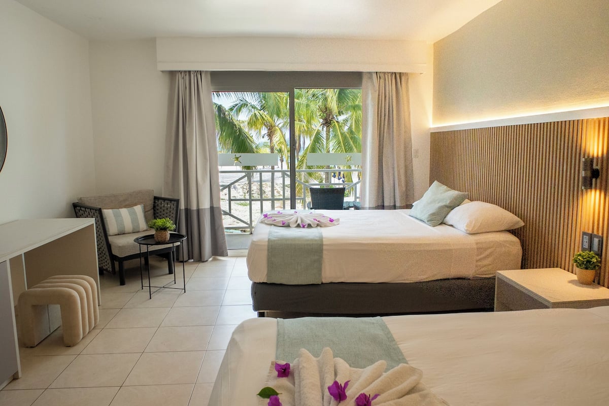 A hotel room with a bed, a desk, a chair, and a balcony with a view of palm trees.