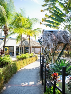 Un sentier bordé de plantes tropicales et de palmiers, qui mène à un bâtiment doté d’une toiture en chaume.