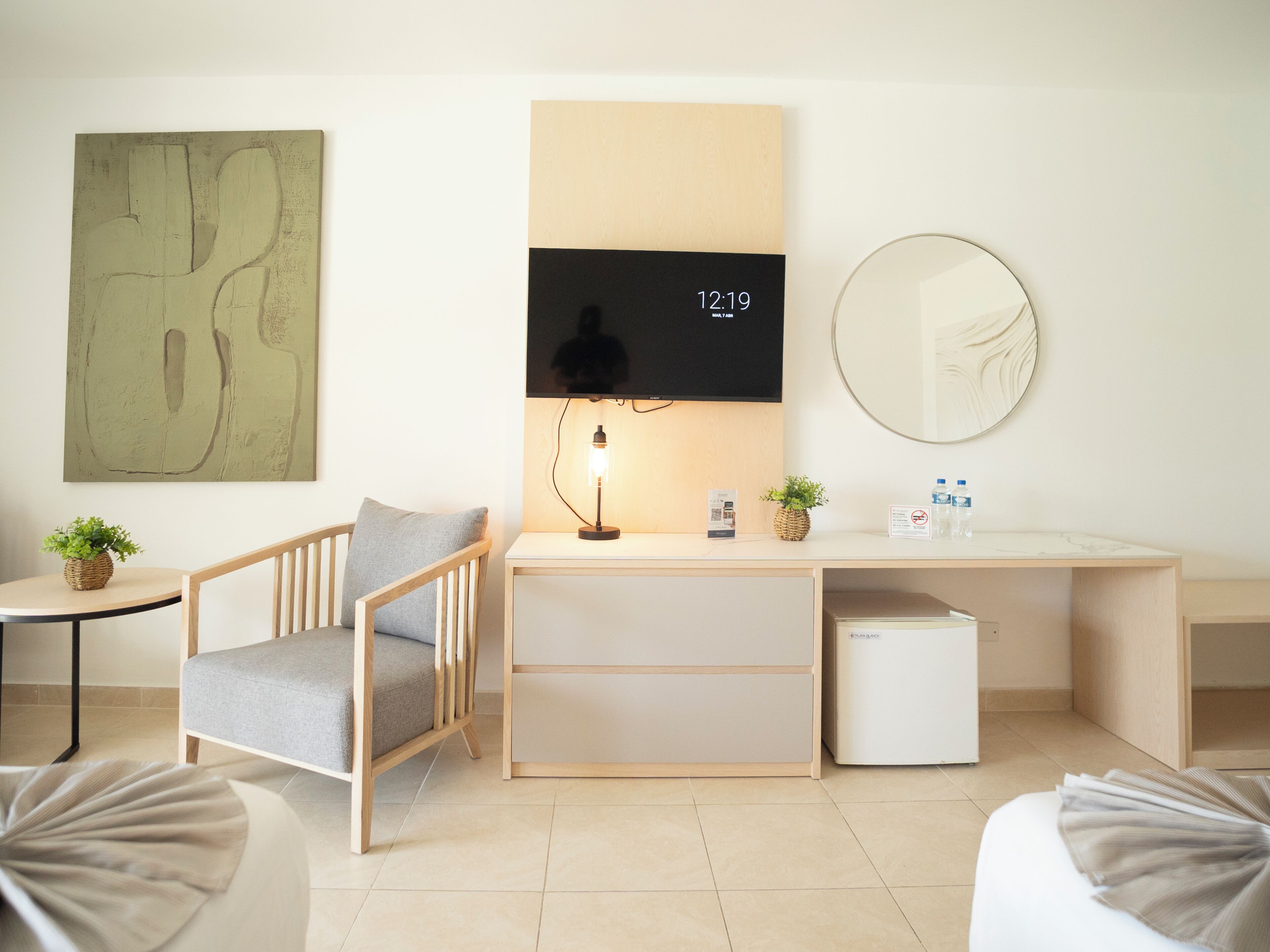 A modern hotel room with a desk, chair, small fridge, and a flat-screen TV displaying the time.