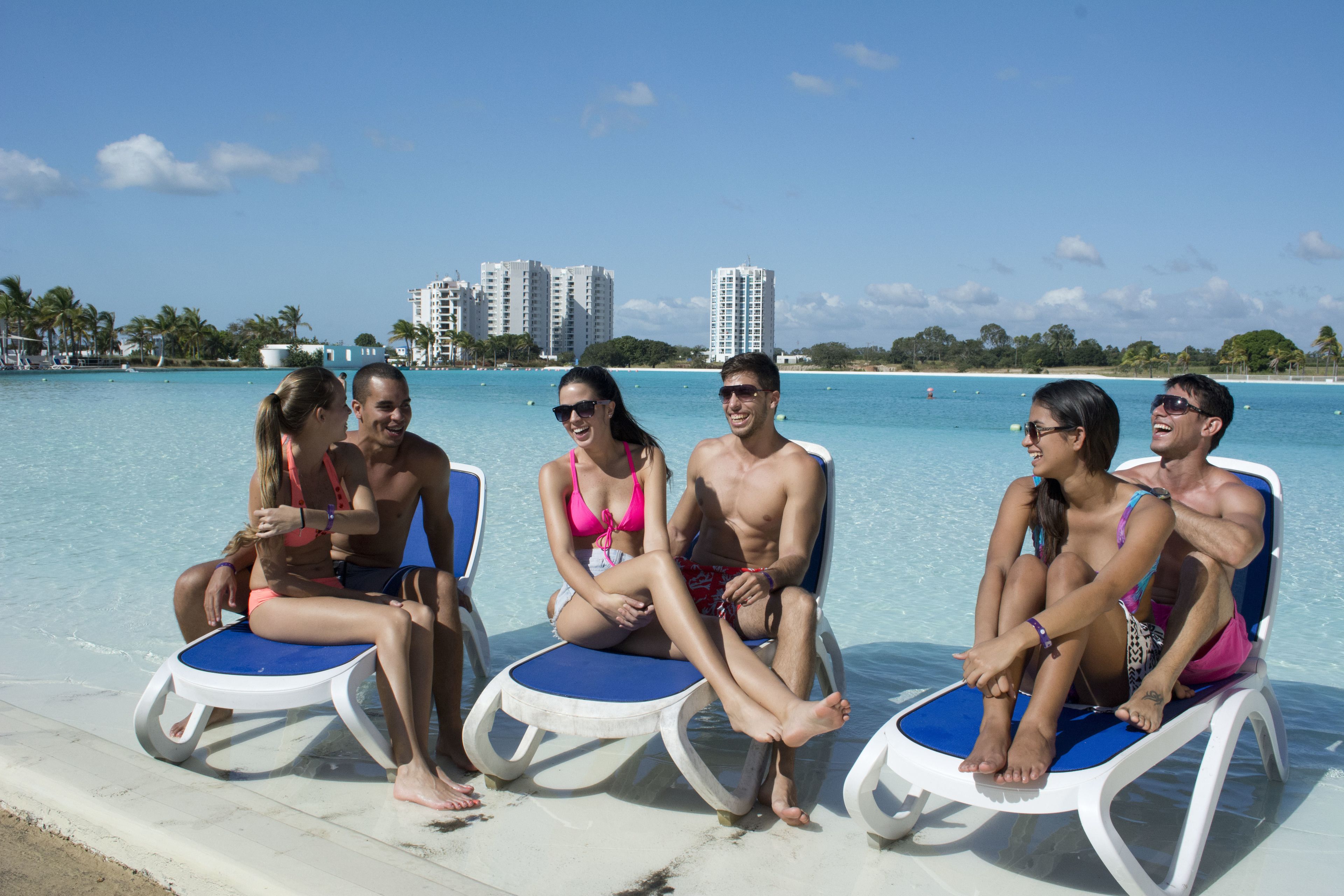 Un grupo de personas sentadas en sillones de descanso junto a una piscina, con vista clara al océano y edificios al fondo.