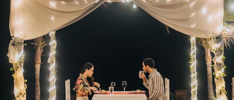 A couple seated at a table under a canopy with string lights, surrounded by palm trees and a wooden deck.