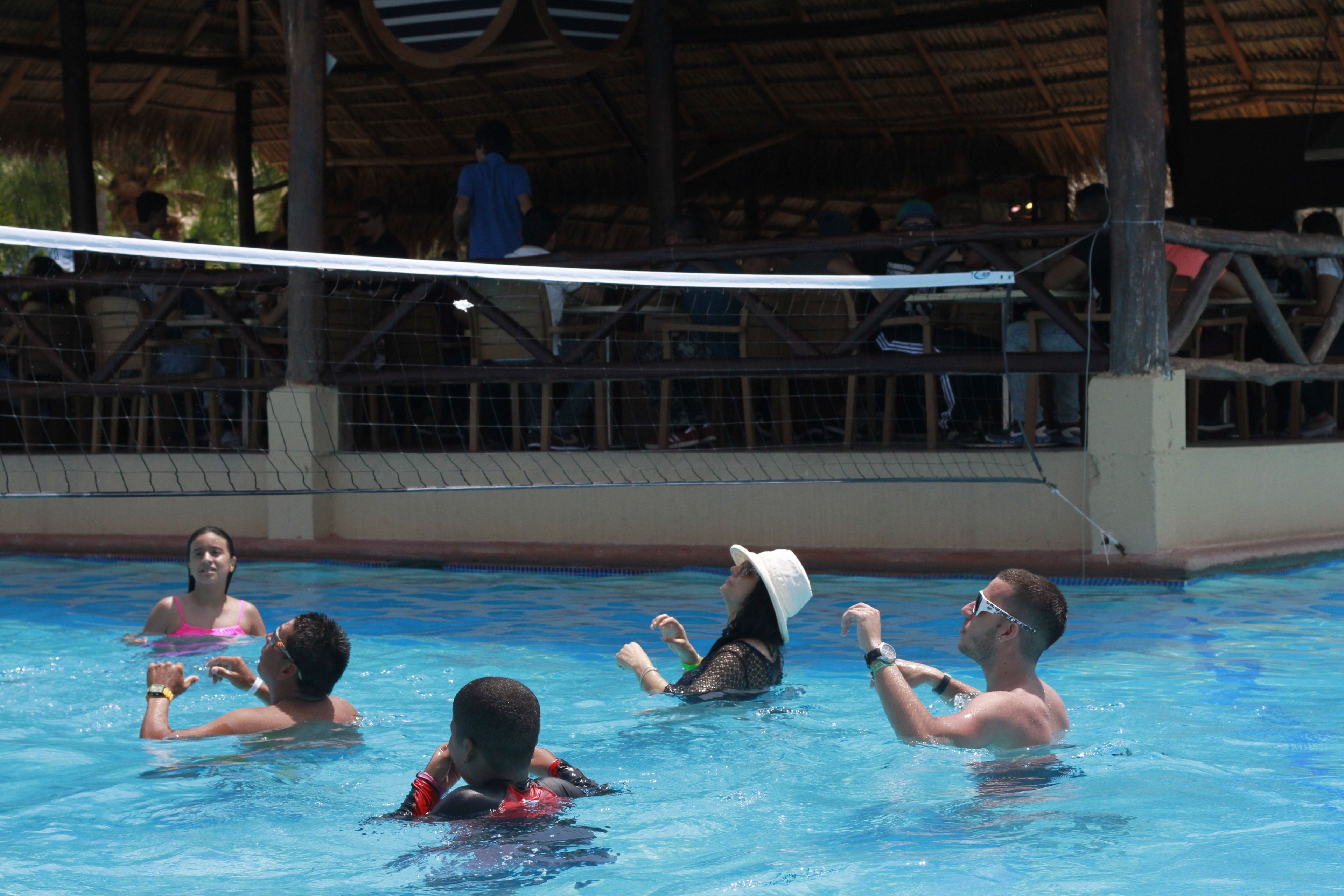 A swimming pool with people in the water, a volleyball net, and a covered spectator area.