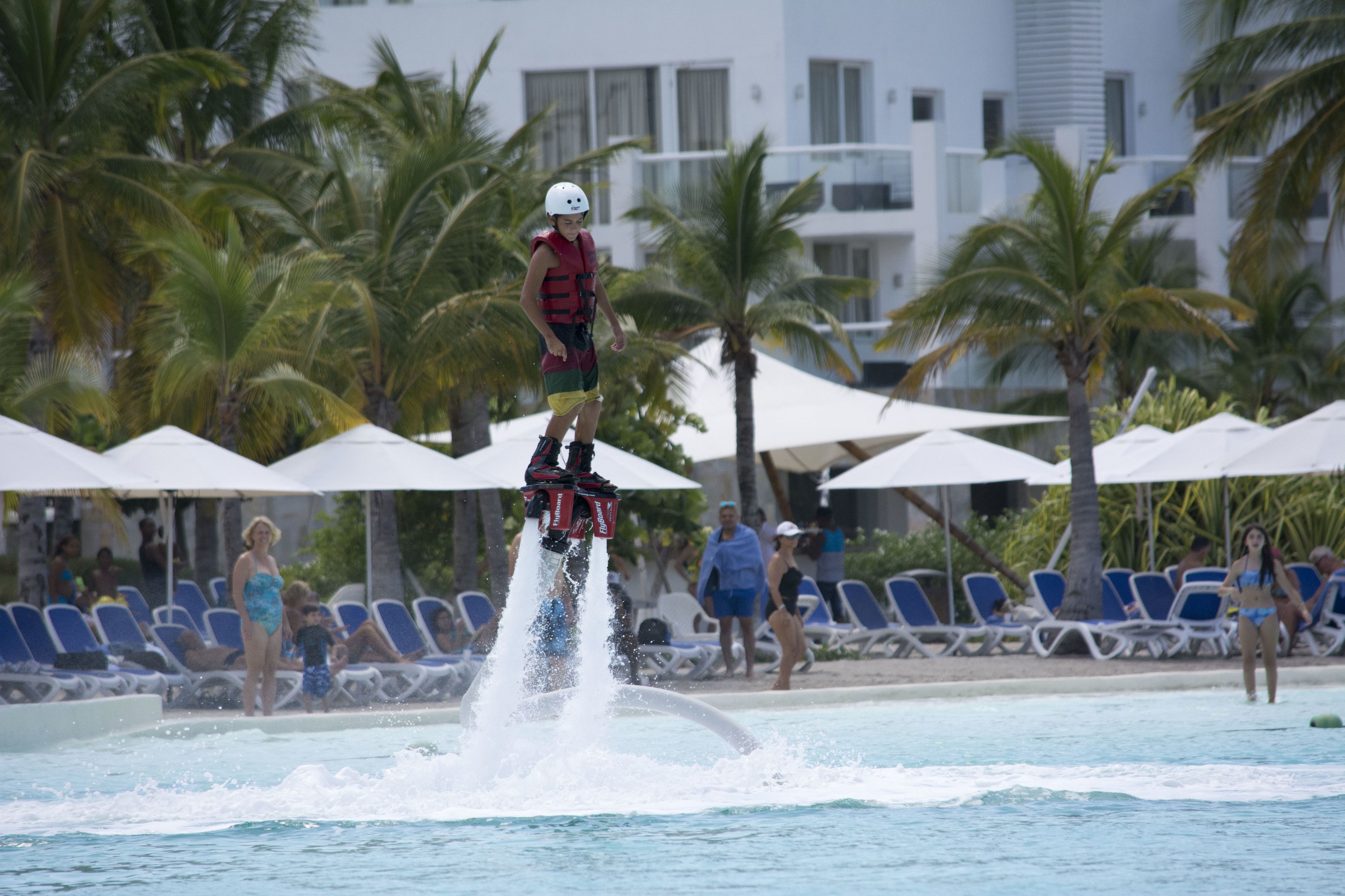 A person performing a water stunt at a pool with palm trees and lounge chairs in the background.