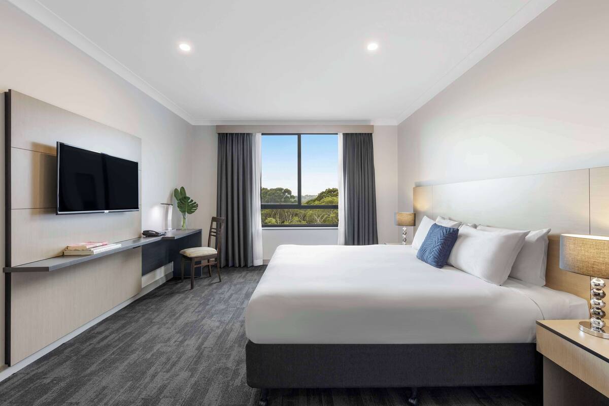 A modern hotel room with a large bed, a flat-screen TV, a desk, and a window with a view of greenery.