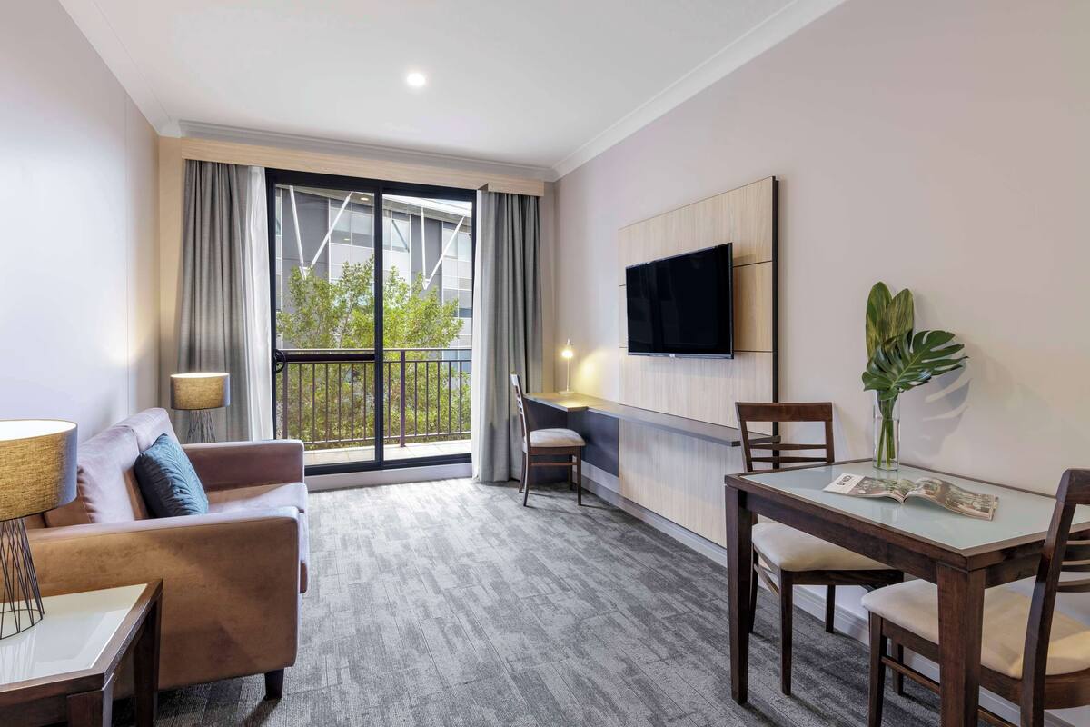A modern hotel room with a sofa, a dining table, a TV mounted on the wall, and a balcony with curtains.
